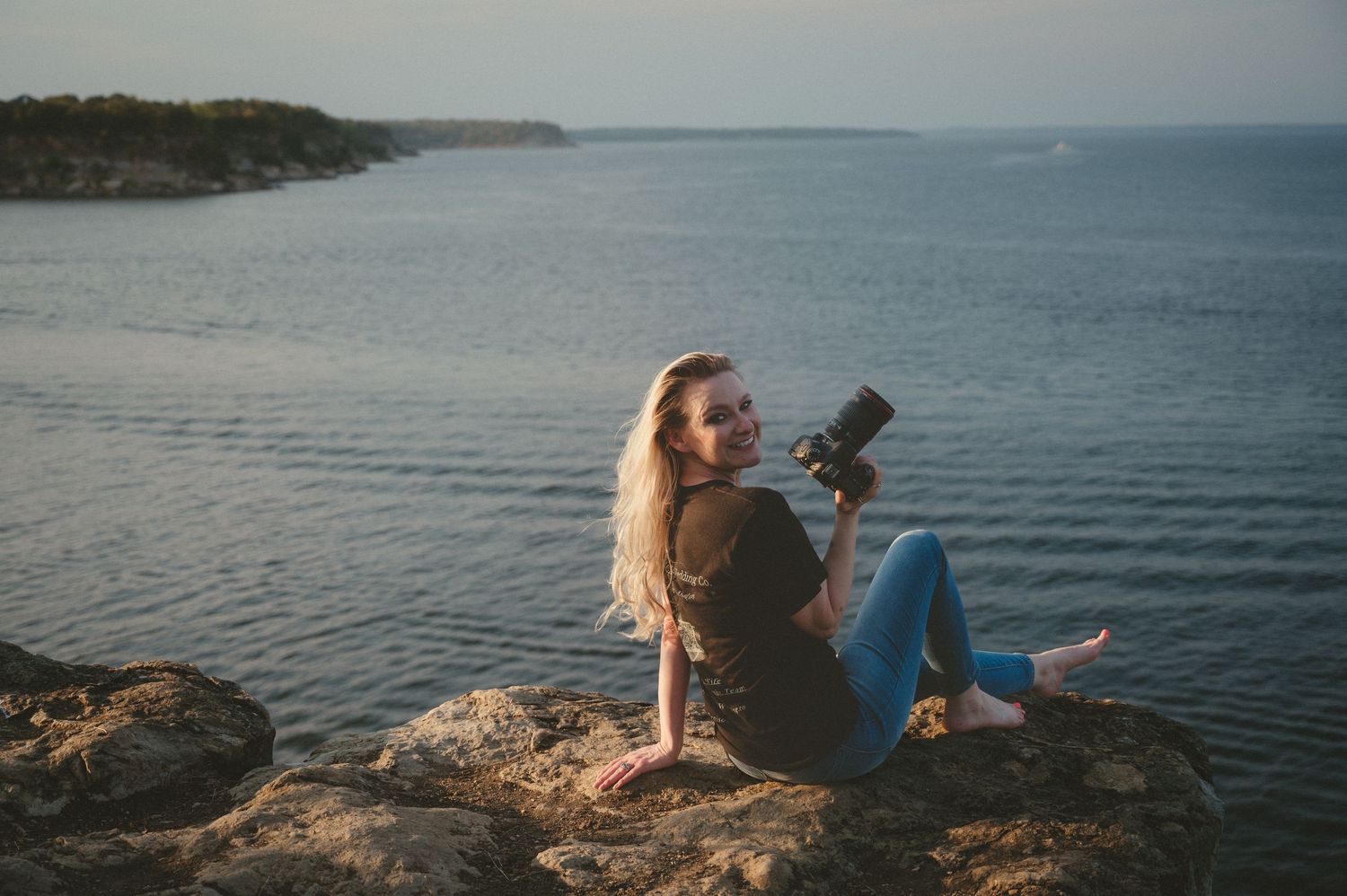 Silhouetted figure stands at edge of tranquil lake during golden sunset with rocky shoreline in view.