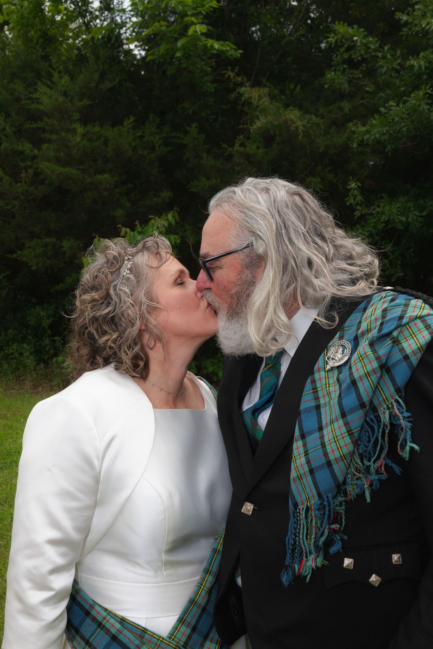 A romantic wedding moment captured outdoors with the couple wearing coordinating white, green, and blue tartan Scottish attire.