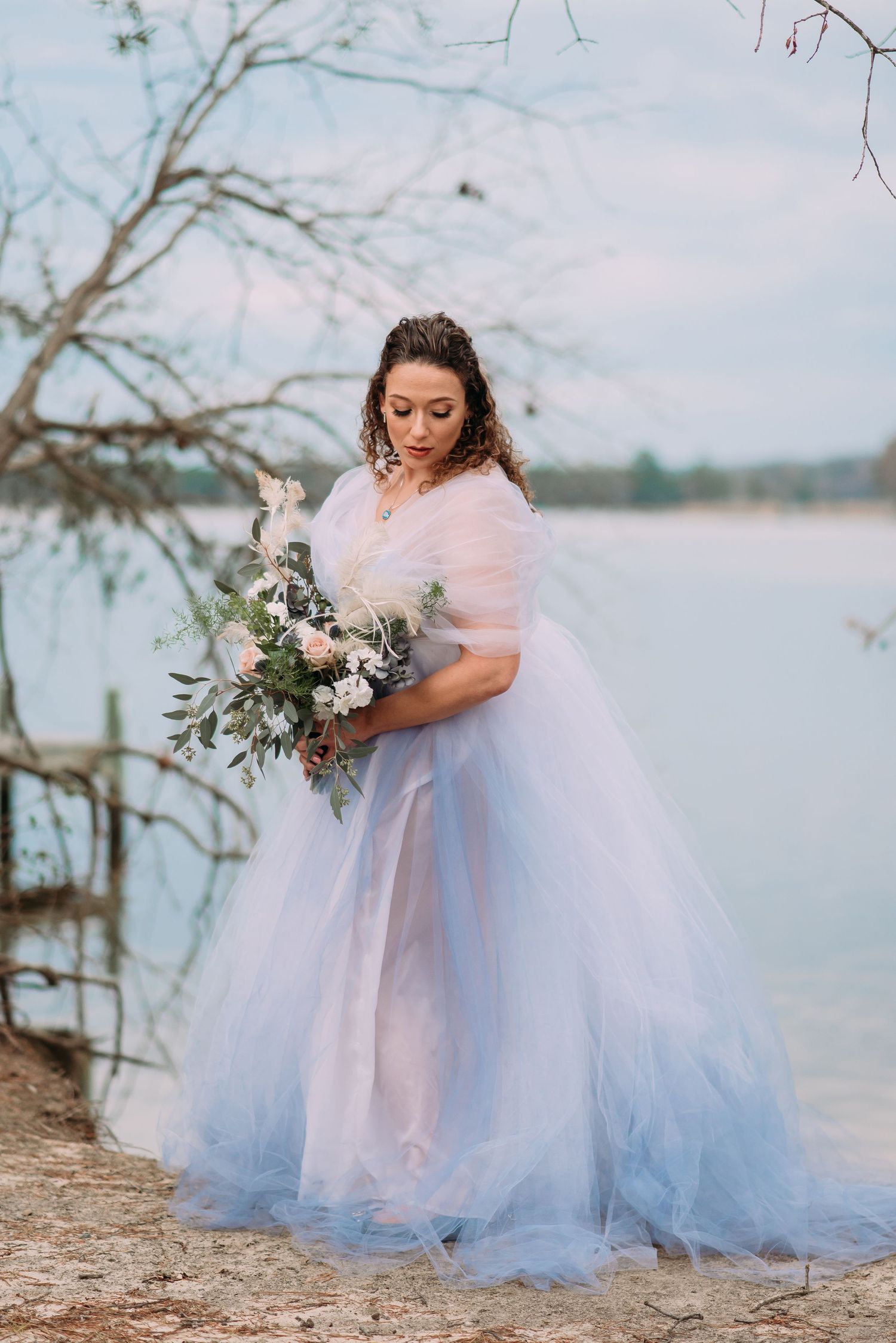 Bride in blue tulle gown holding floral bouquet by lakeside with bare trees in soft natural light.