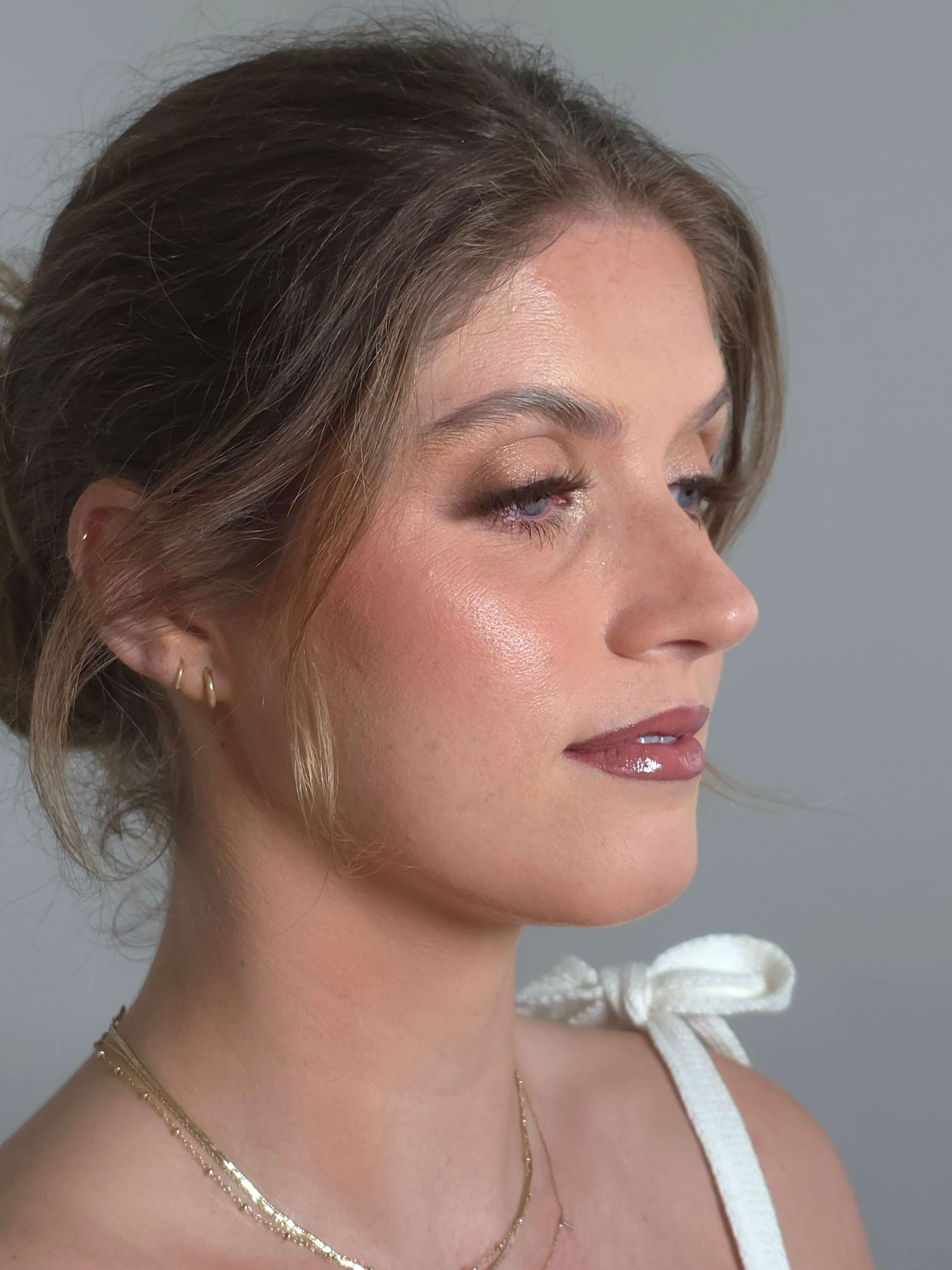 Woman with elegant updo, glowing skin, burgundy lips, and gold necklace wearing a white bow top.