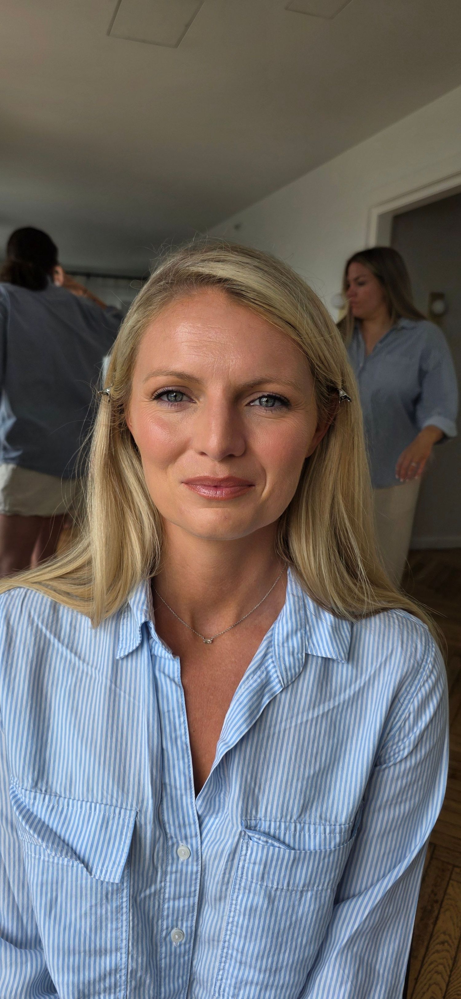 Blonde woman in blue striped shirt smiling indoors with people in background.