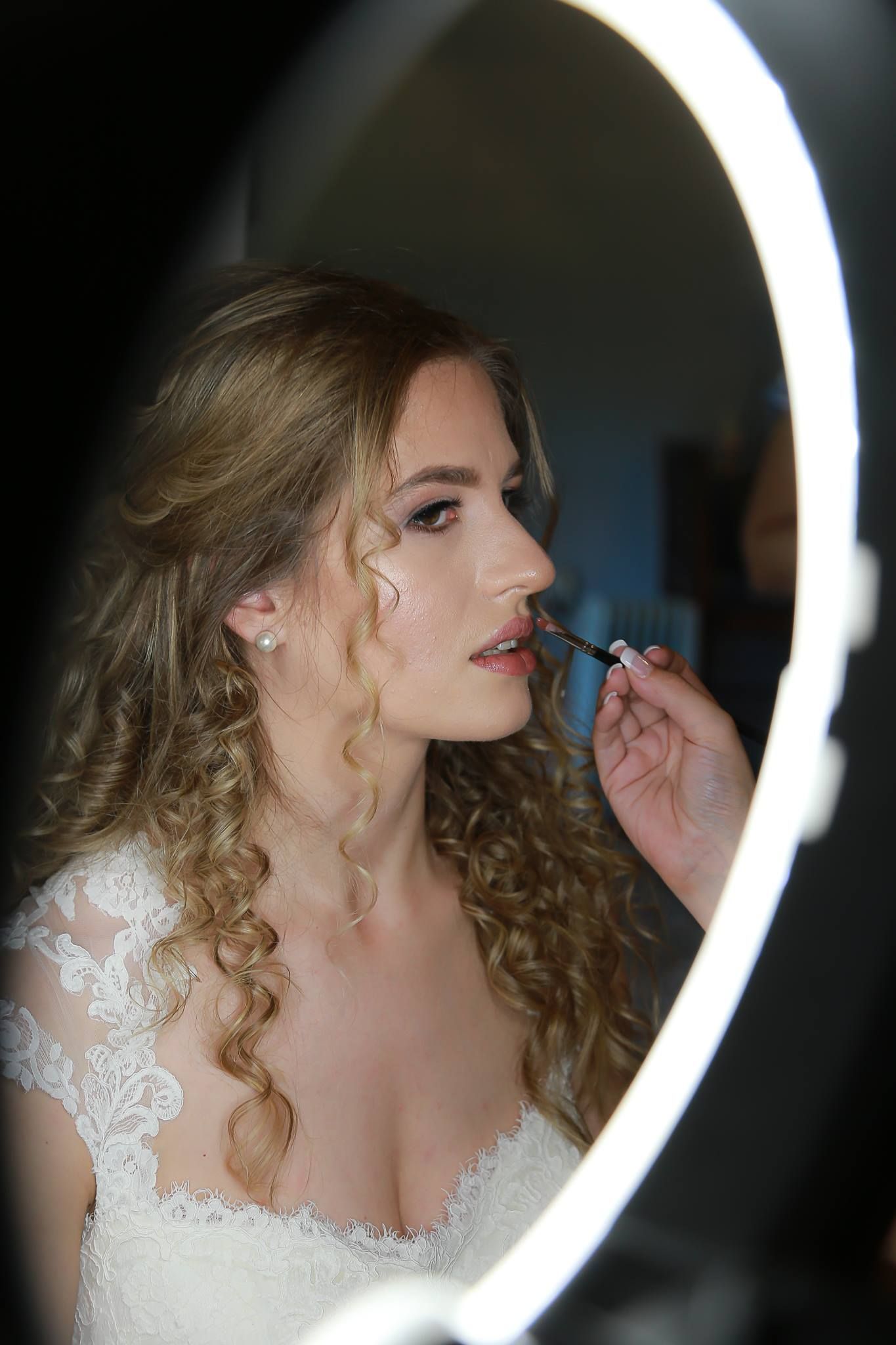 Bride in white lace dress getting lip makeup applied by artist, reflected in illuminated oval mirror.