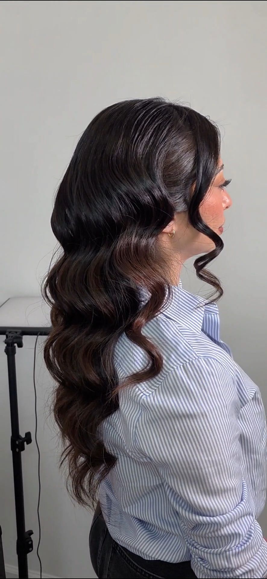 Woman with long dark brown wavy hair styled in glamorous Hollywood waves, wearing a blue striped shirt.
