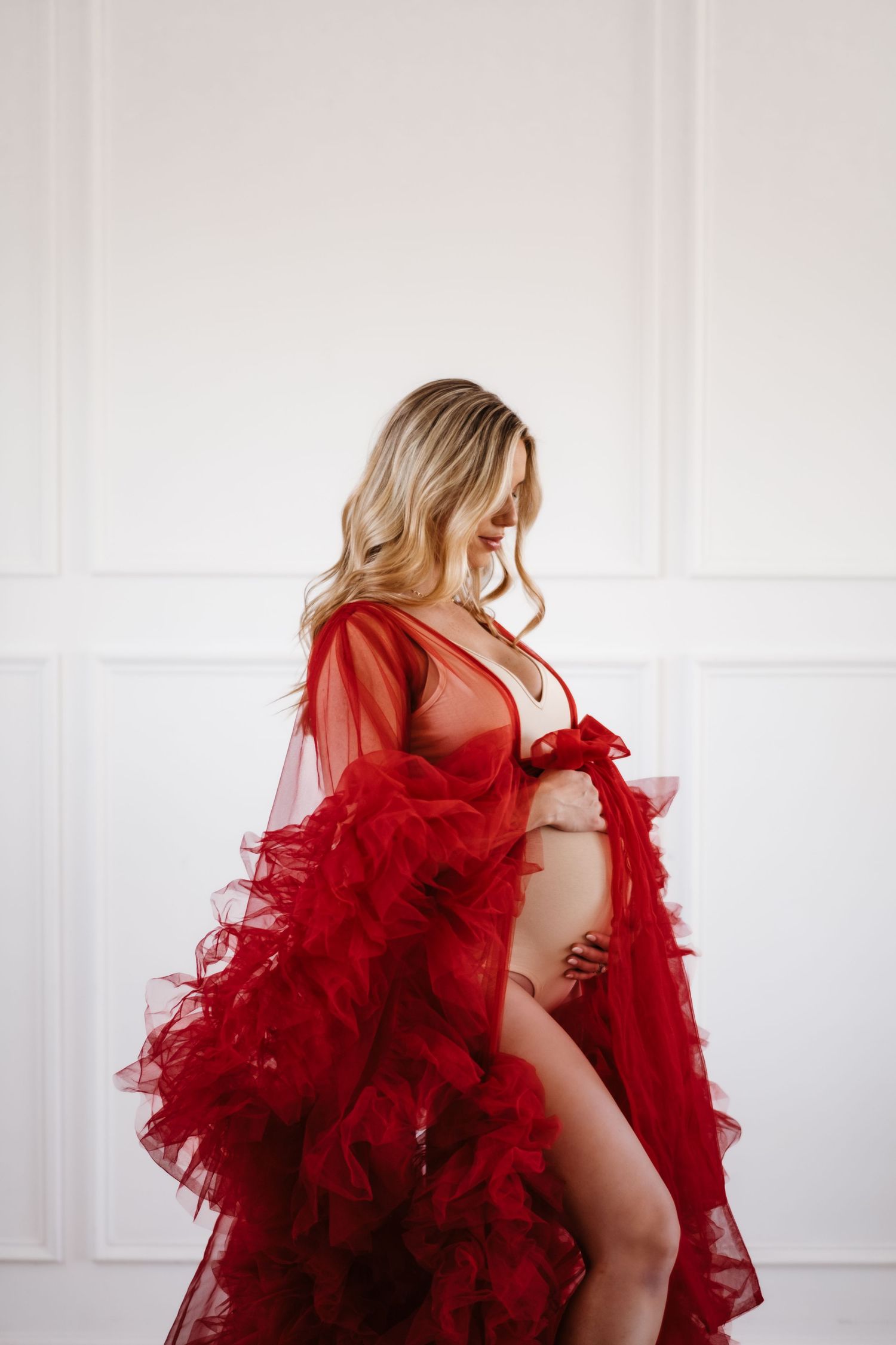 A maternity boudoir photo featuring a flowing red tulle robe against a white wall.