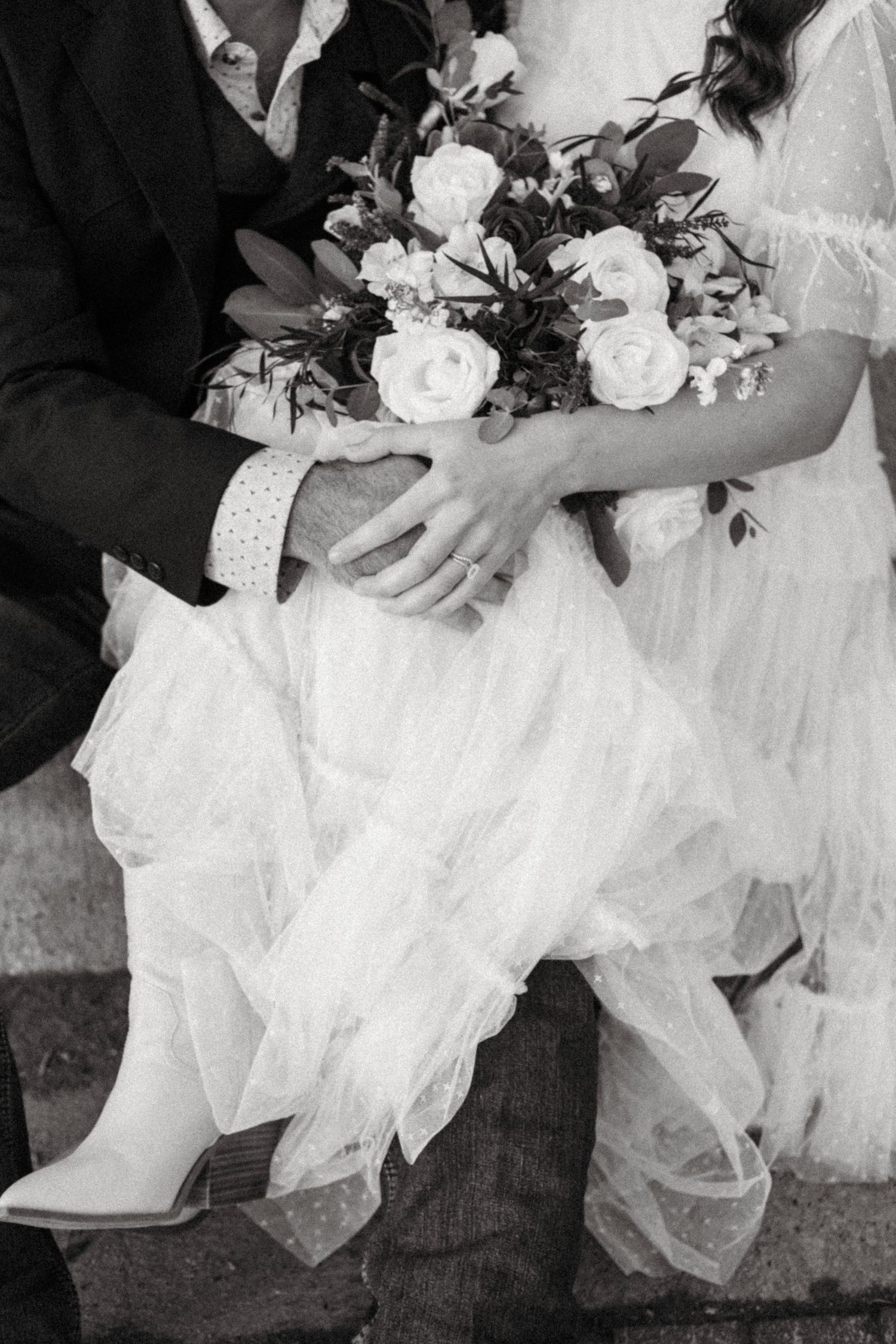 Black and white detail shot of wedding bouquet with white roses and flowing dress fabric.