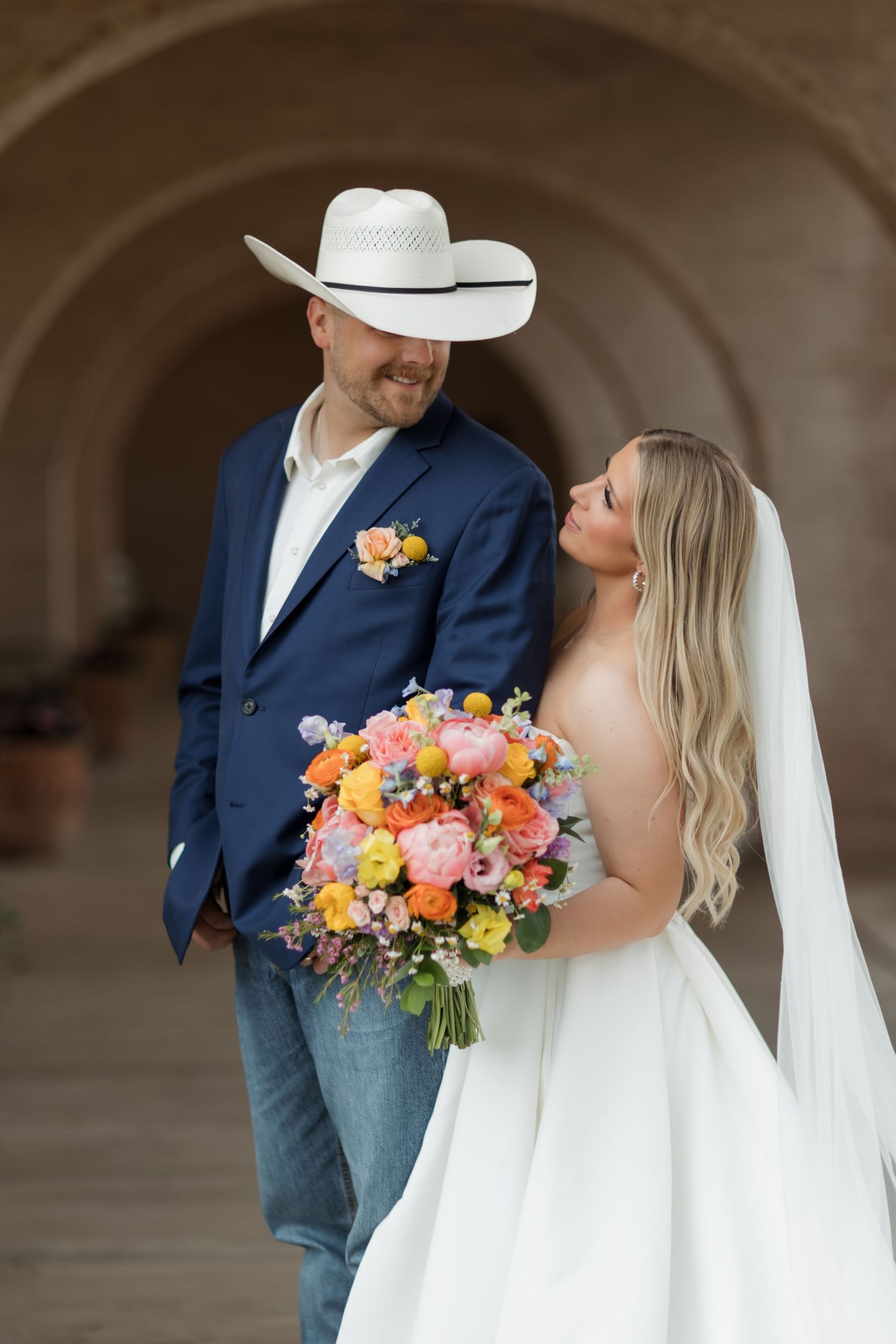 Bride and groom pose under stone arches, she holds colorful bouquet in wedding dress, he wears navy suit and white cowboy hat.