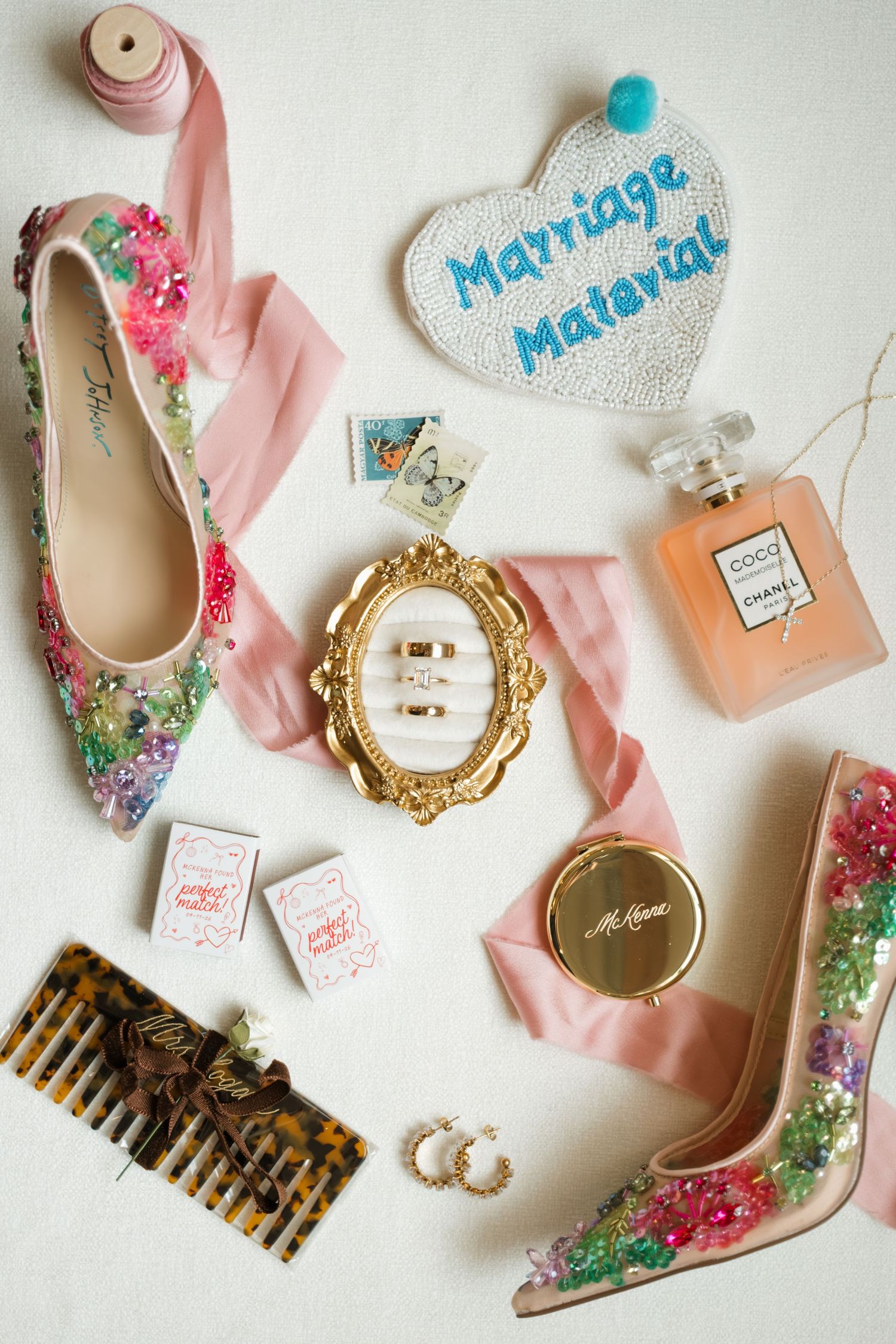 Flat lay of floral embellished bridal shoes, Coco Chanel perfume, gold mirror, hair comb, and Marriage Material heart patch.