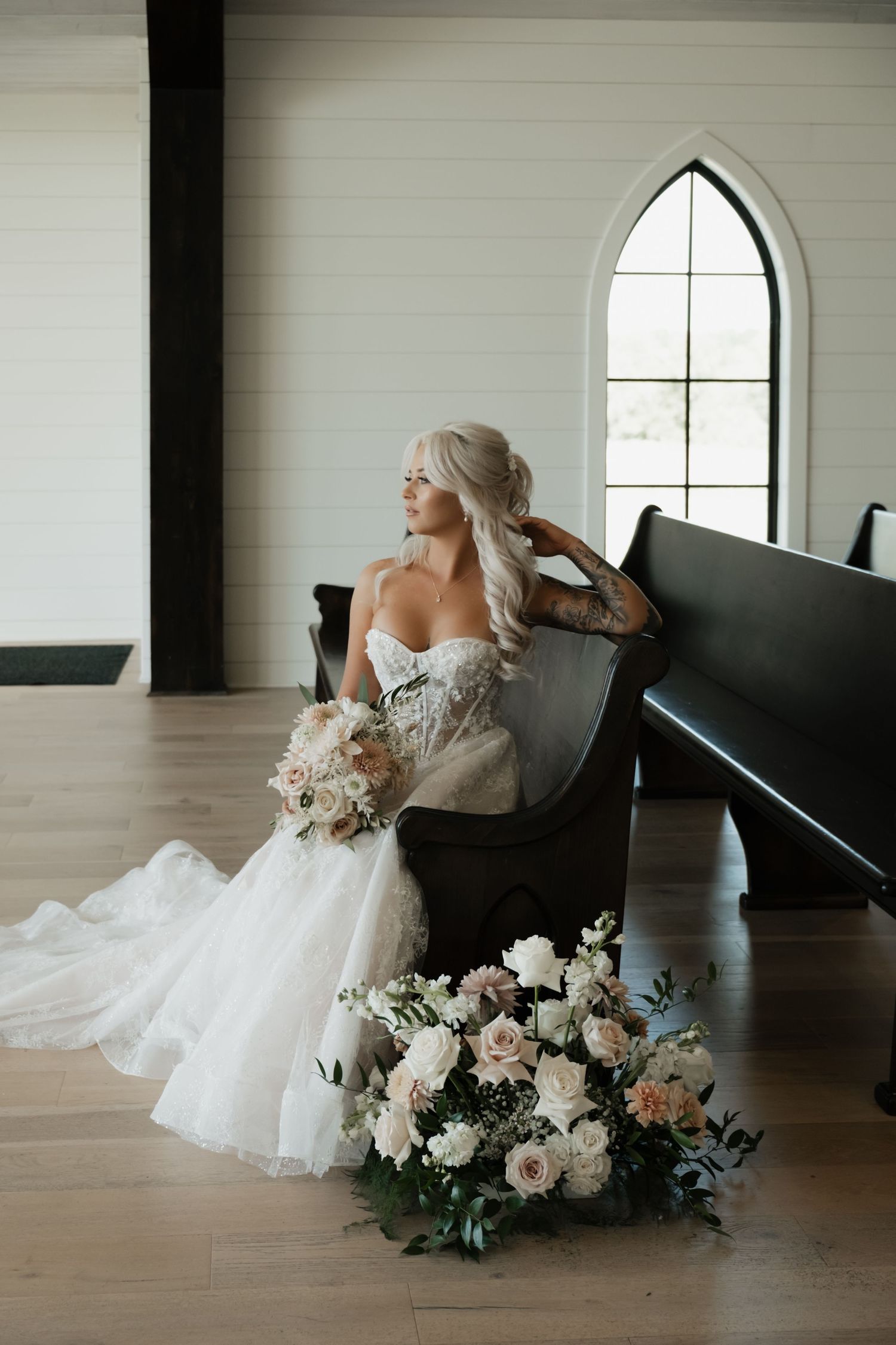 Elegant bride in off-shoulder white gown sits on church pew beside romantic floral arrangement in soft pastel tones.