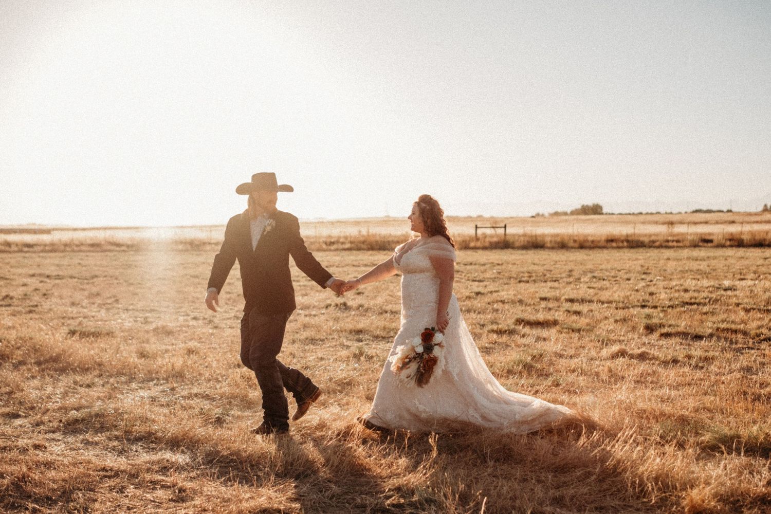 A couple running hand in hand through a sunlit field during their rustic wedding.