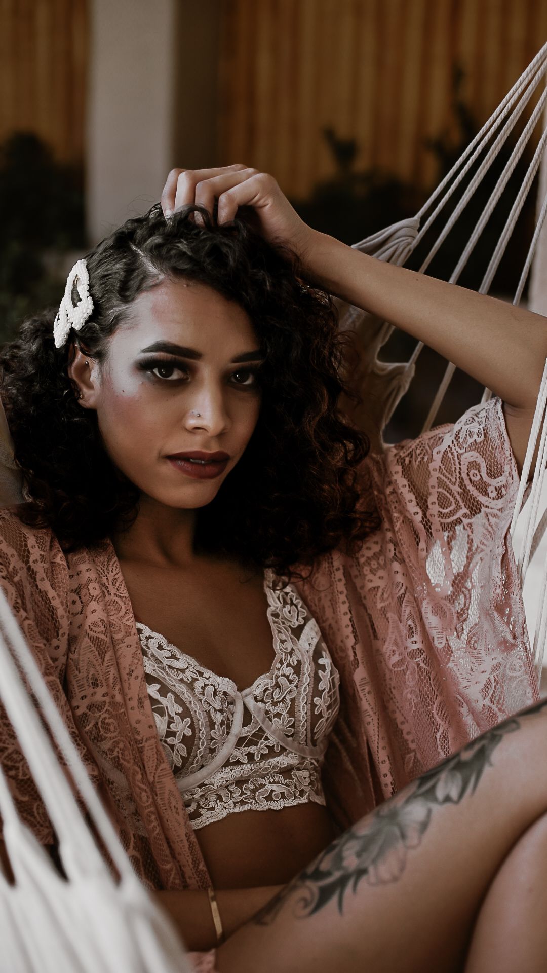 A person in pink lace lingerie poses seductively in a white hammock in a romantic boudoir setting.