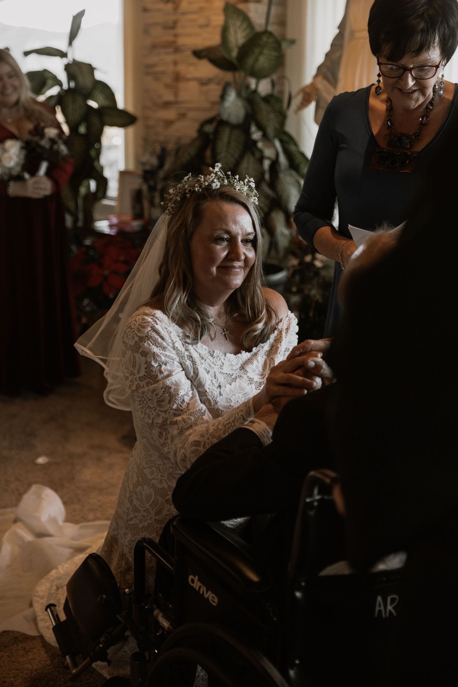 Bride kneels during an intimate indoor elopement ceremony, holding hands with her partner in a wheelchair as they exchange vows.