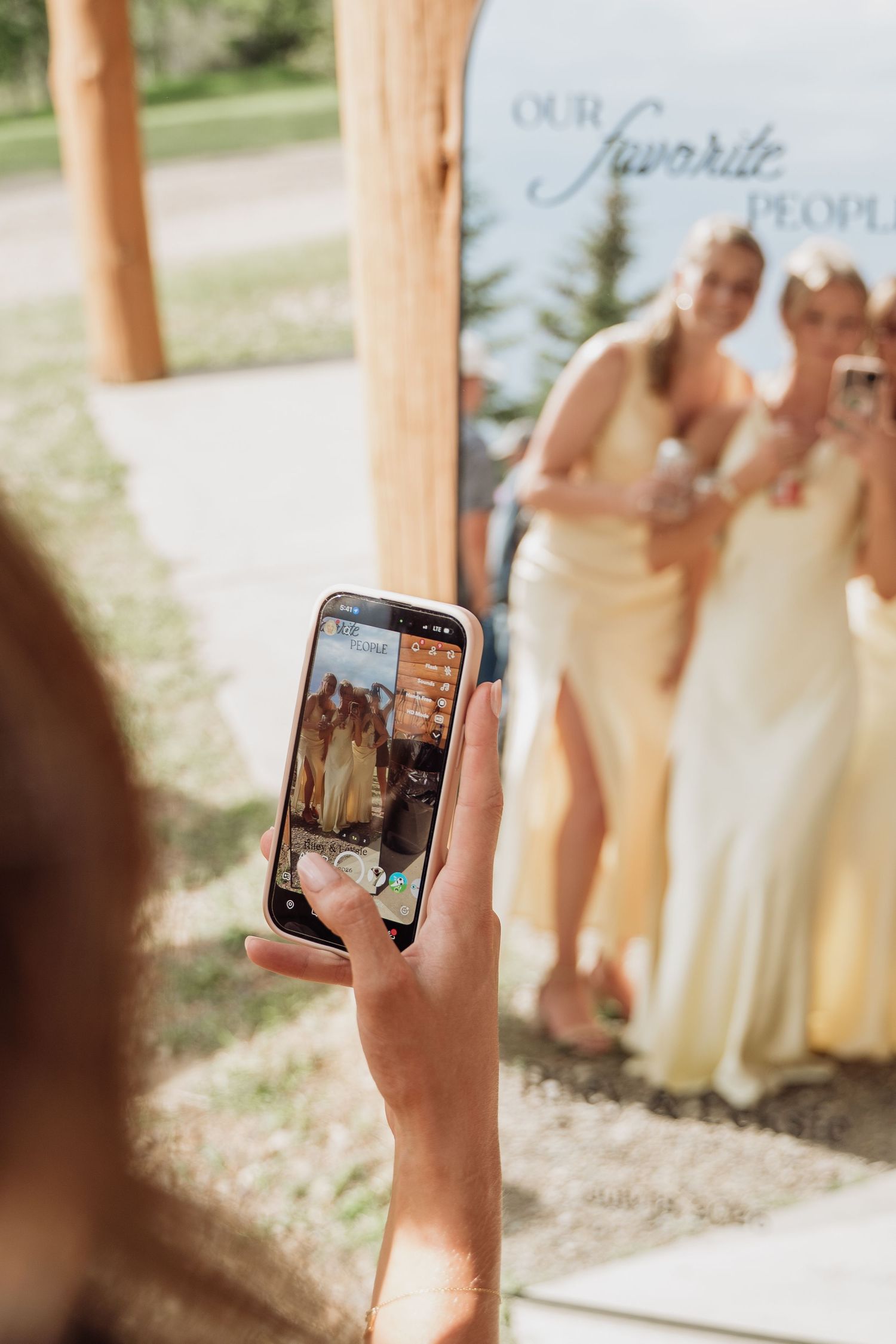 Guest photographing bridesmaids in yellow dresses at outdoor wedding photo booth.