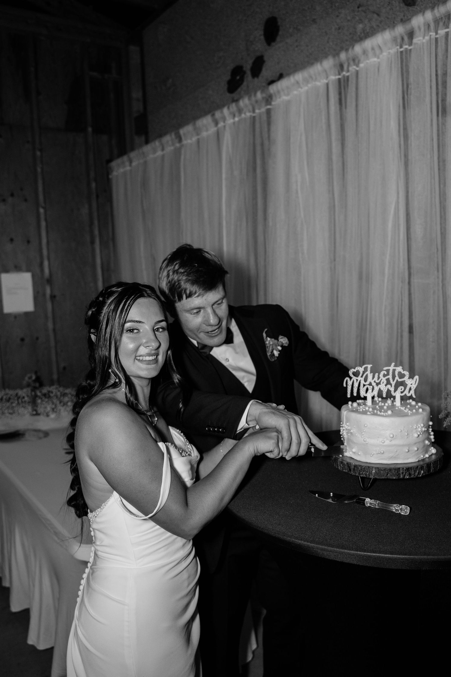 Black and white photo of smiling bride and groom cutting their wedding cake together at reception.