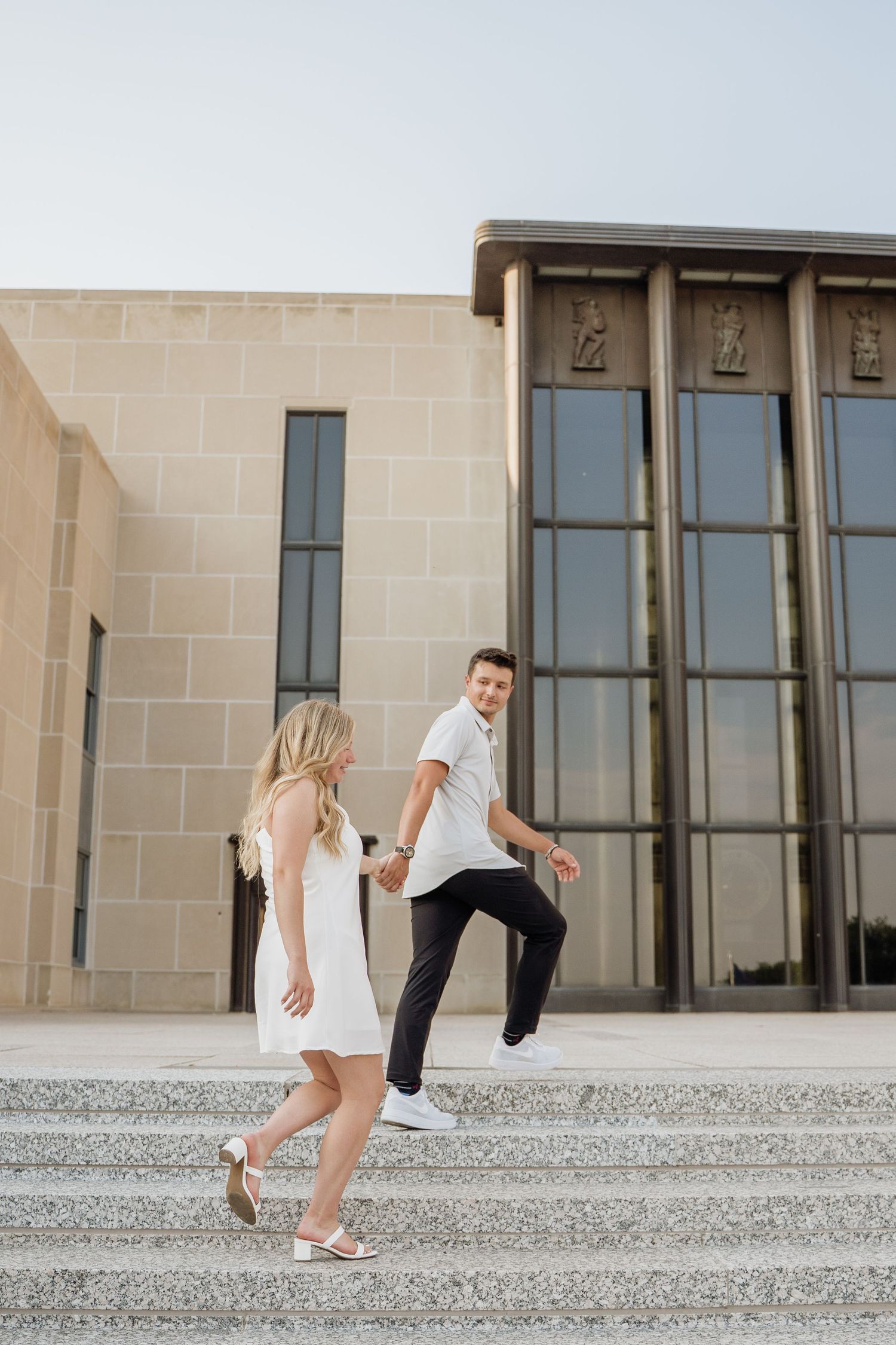 A couple in white outfits walks up outdoor steps in front of a modern building.