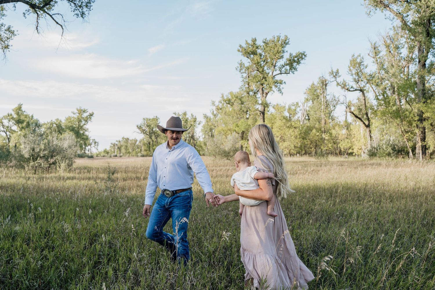A couple walks hand-in-hand through a lush green meadow, man in cowboy hat and woman in flowing dress.