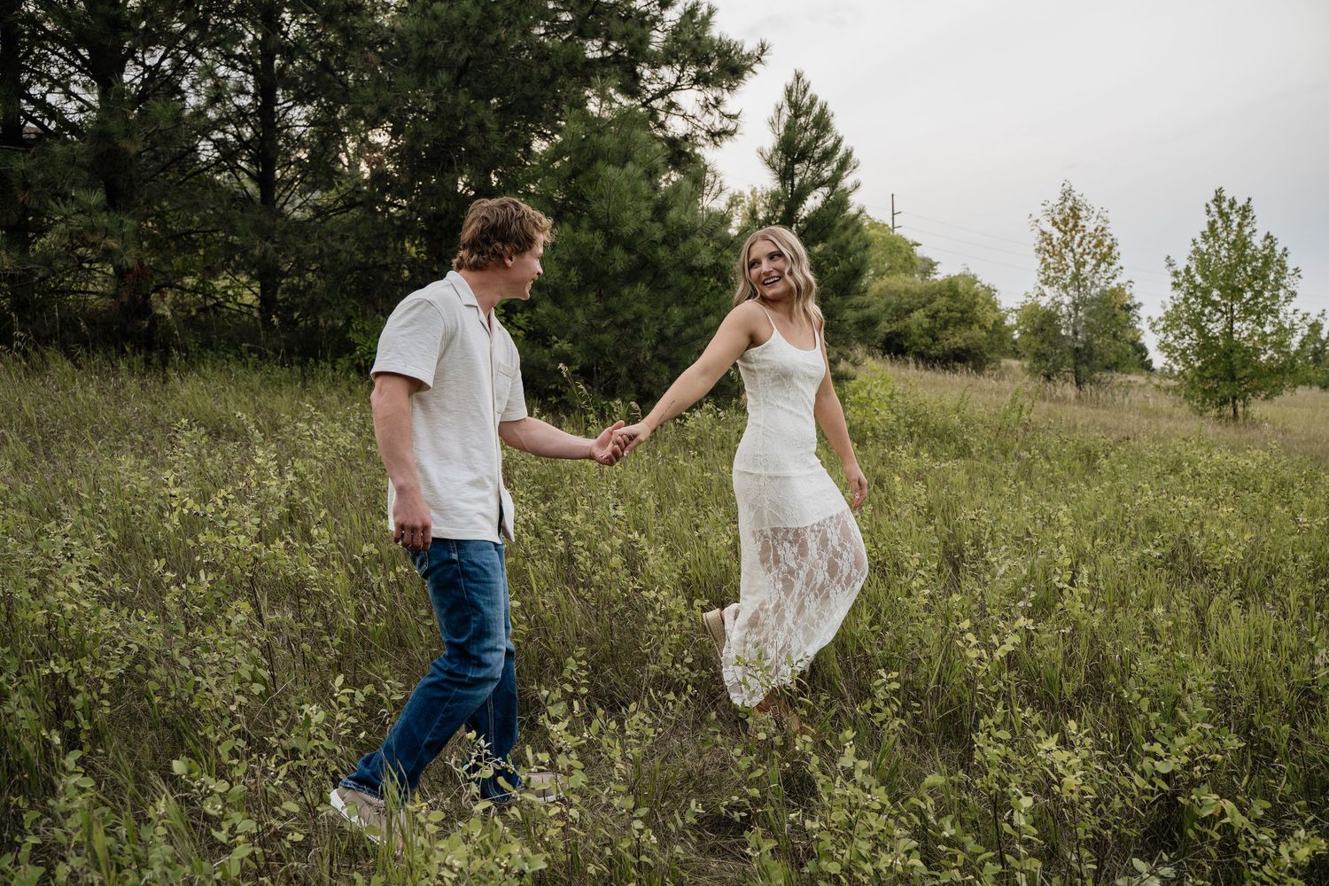 Couple holding hands walking through green meadow, woman in white lace dress, romantic outdoor photo.