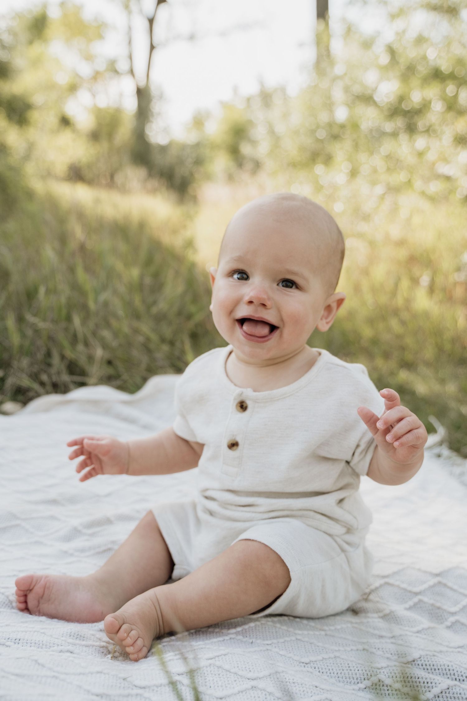 Adorable smiling baby in cream romper sitting on white blanket outdoors in natural setting.