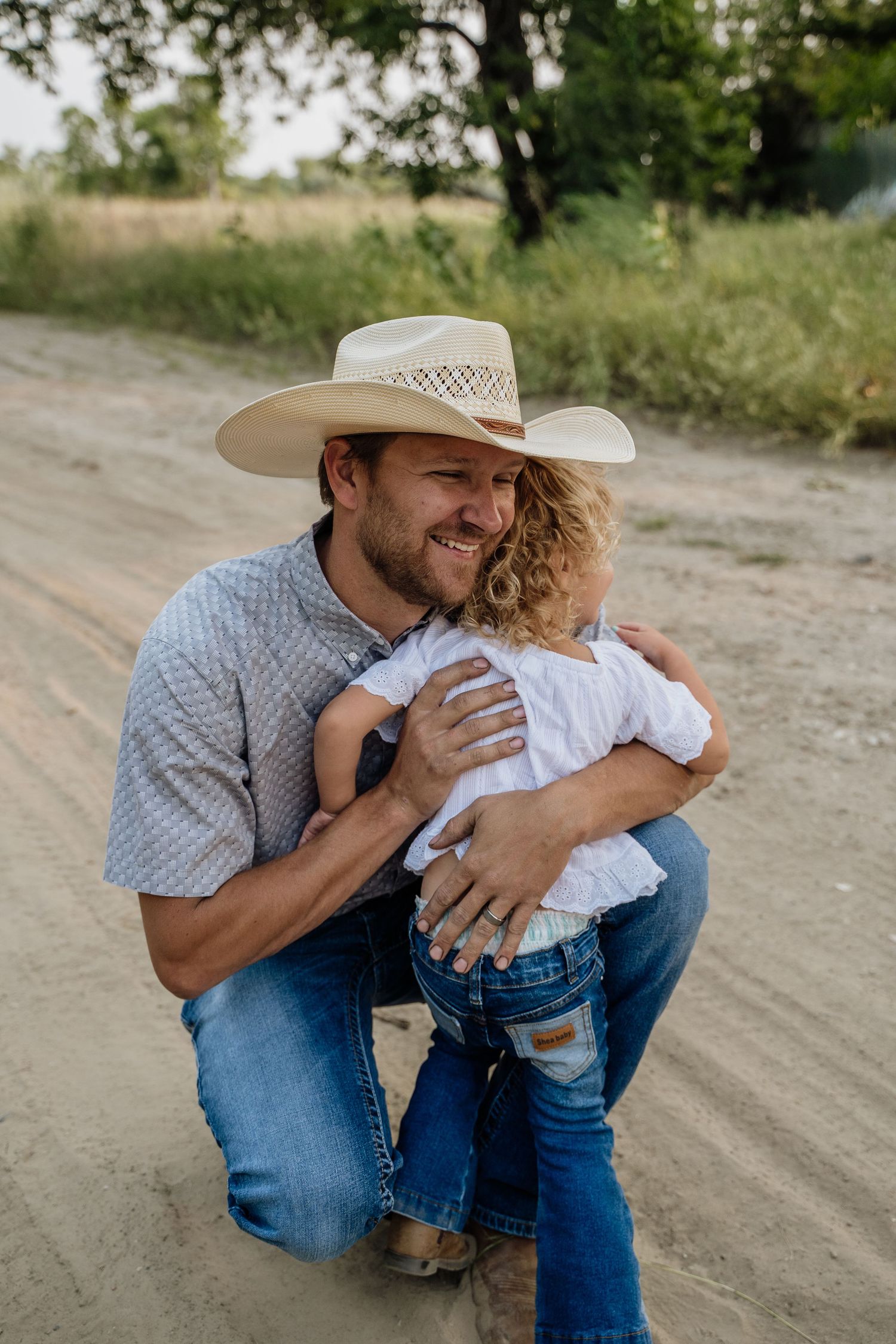 Smiling father in cowboy hat kneeling on dirt road hugging young curly-haired toddler.