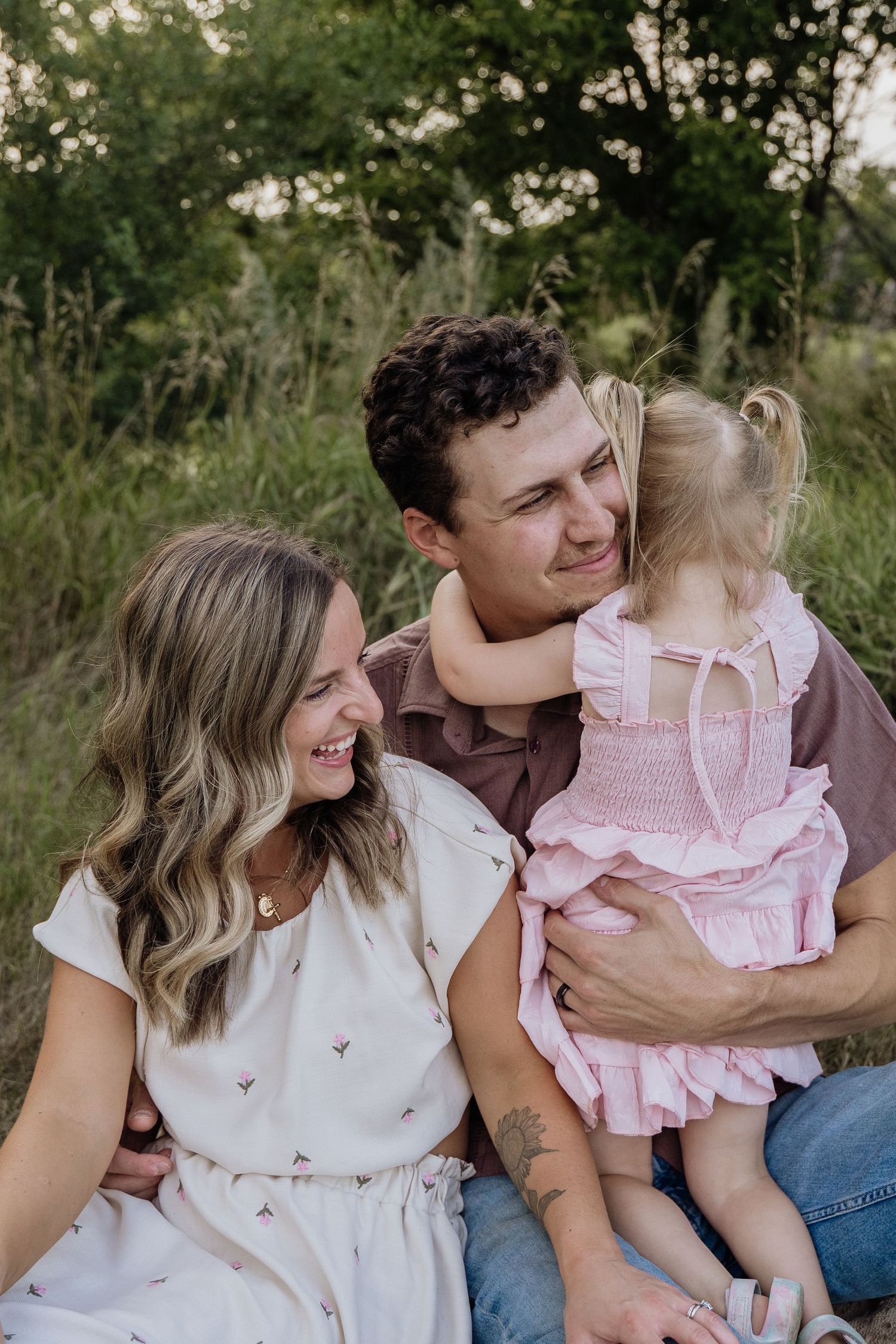 Happy family sitting in grass with curly-haired toddler in pink dress hugging father.