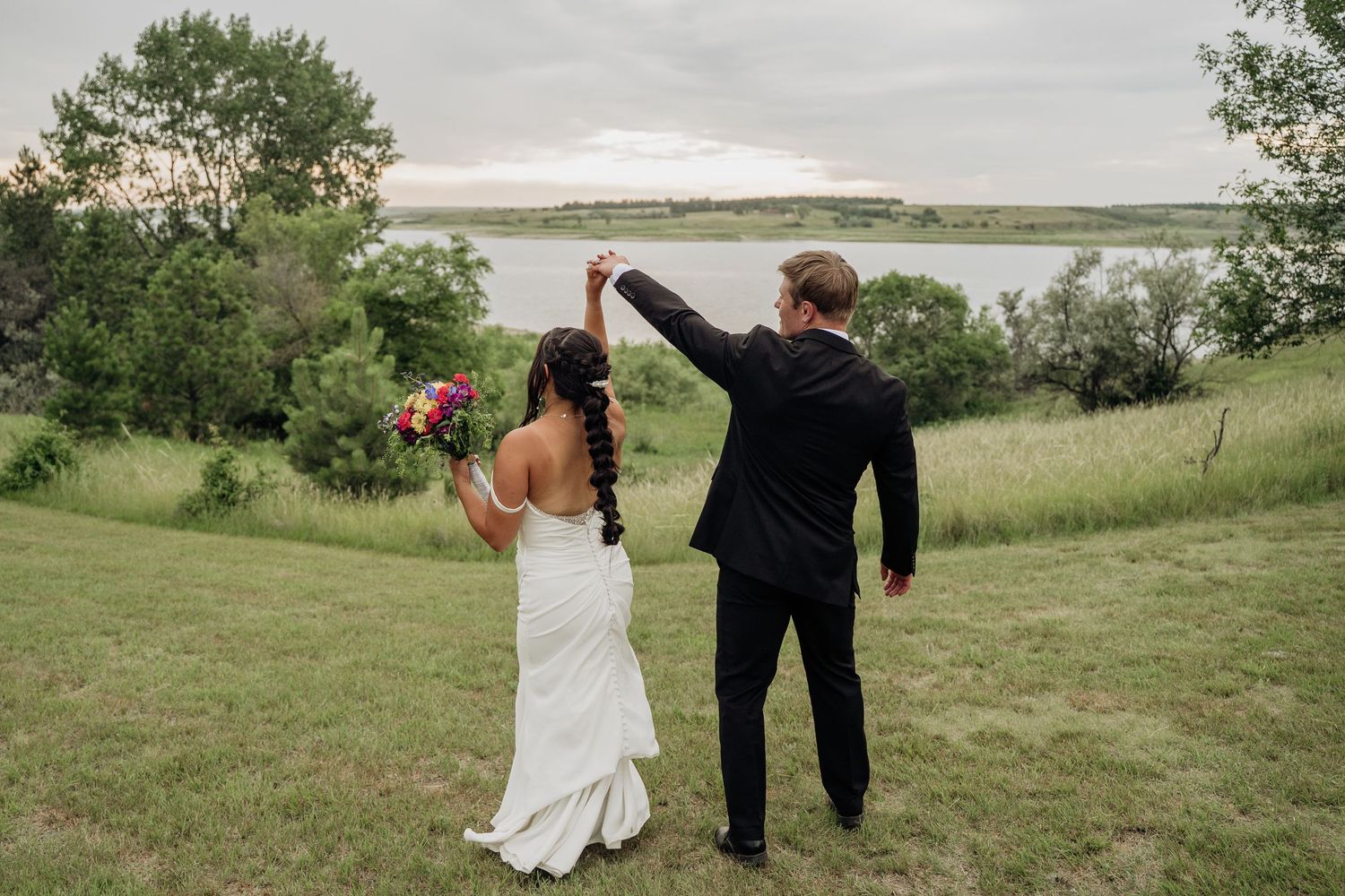 Bride and groom dancing outdoors near a lake at sunset, bride holding a colorful bouquet in a white gown.