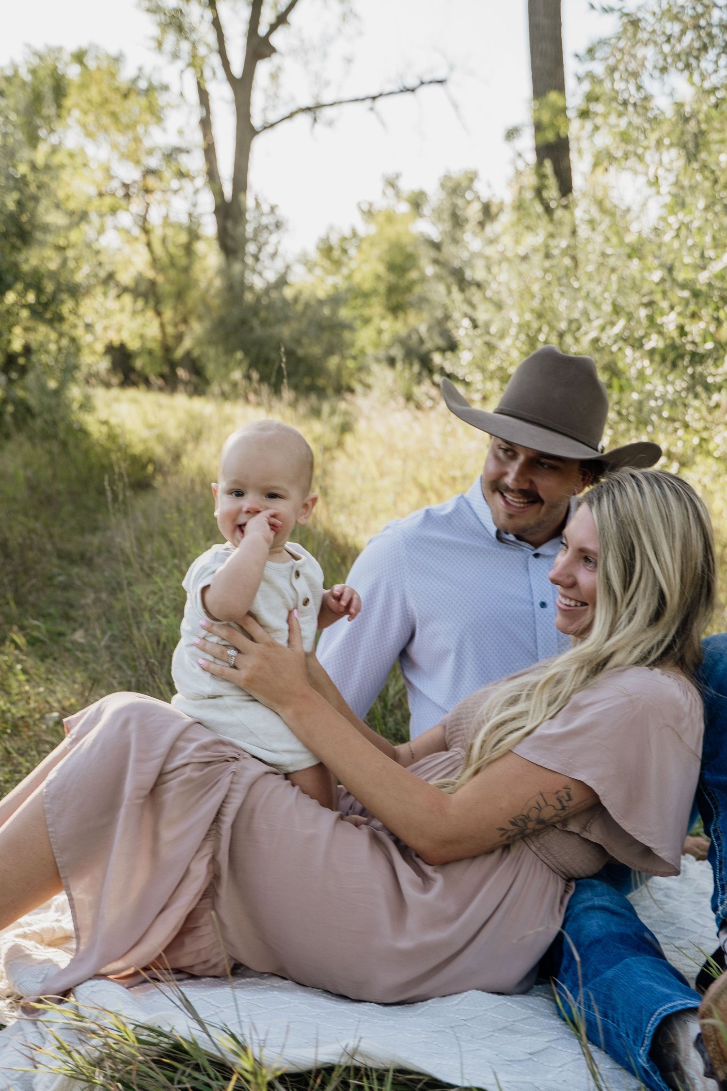 A young family sits outdoors on a blanket, with a smiling man in a cowboy hat and woman holding their happy baby.