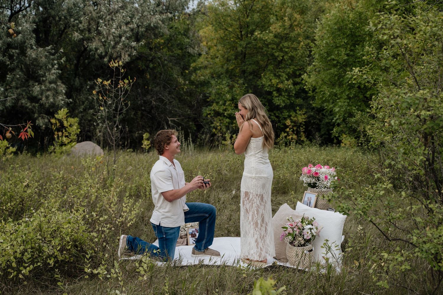 Man proposing to woman outdoors in a meadow with floral decorations and lush green trees in the background.