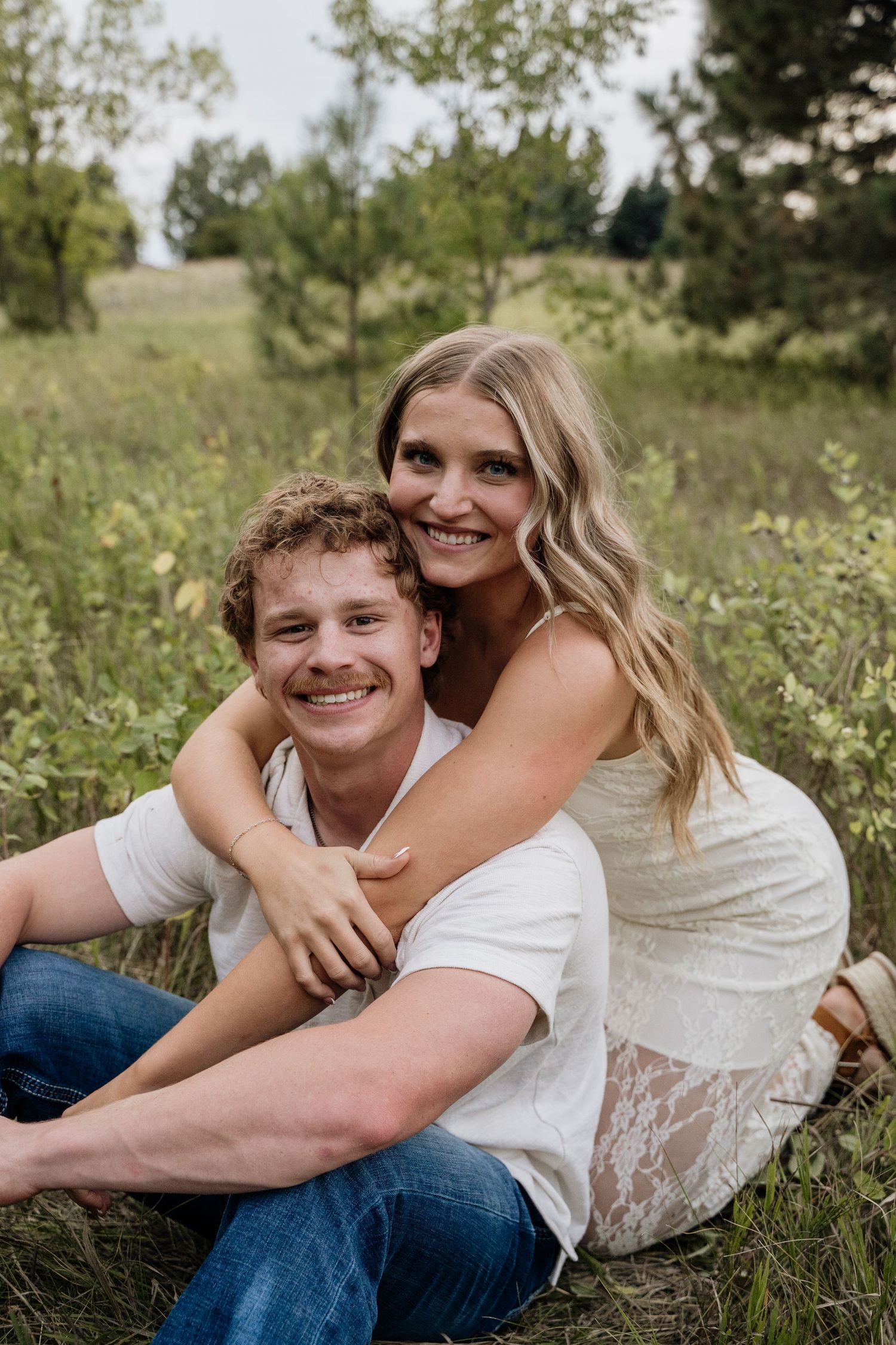 Smiling couple sitting in grass field, woman in white lace dress embracing man from behind, outdoors.