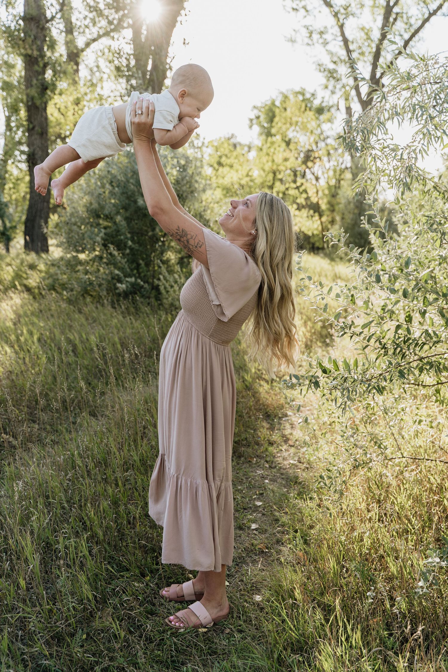 Blonde mother in beige dress lifting laughing baby overhead in sunlit green meadow.