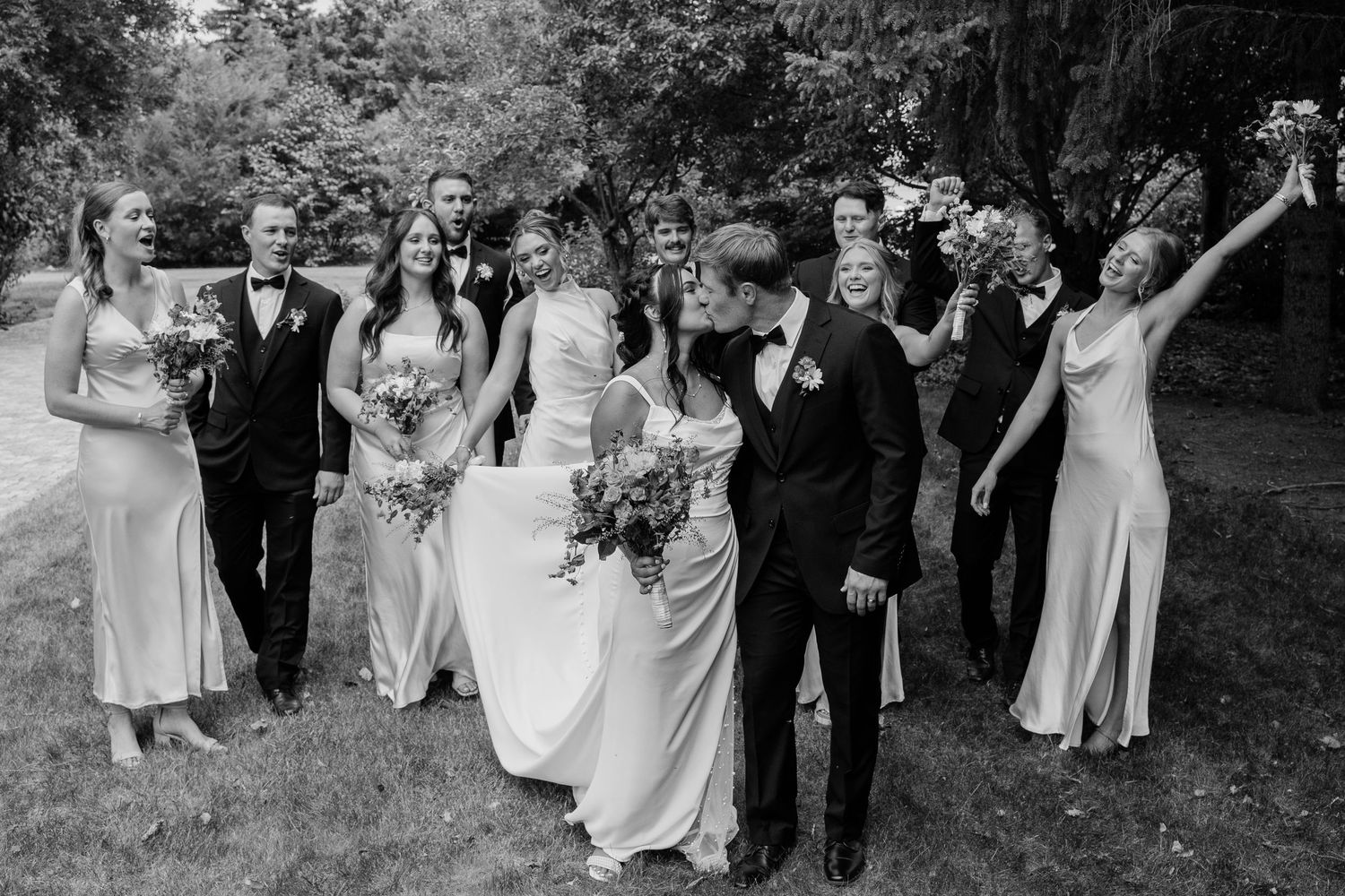 Black and white photo of wedding party celebrating as bride and groom share a kiss outdoors.