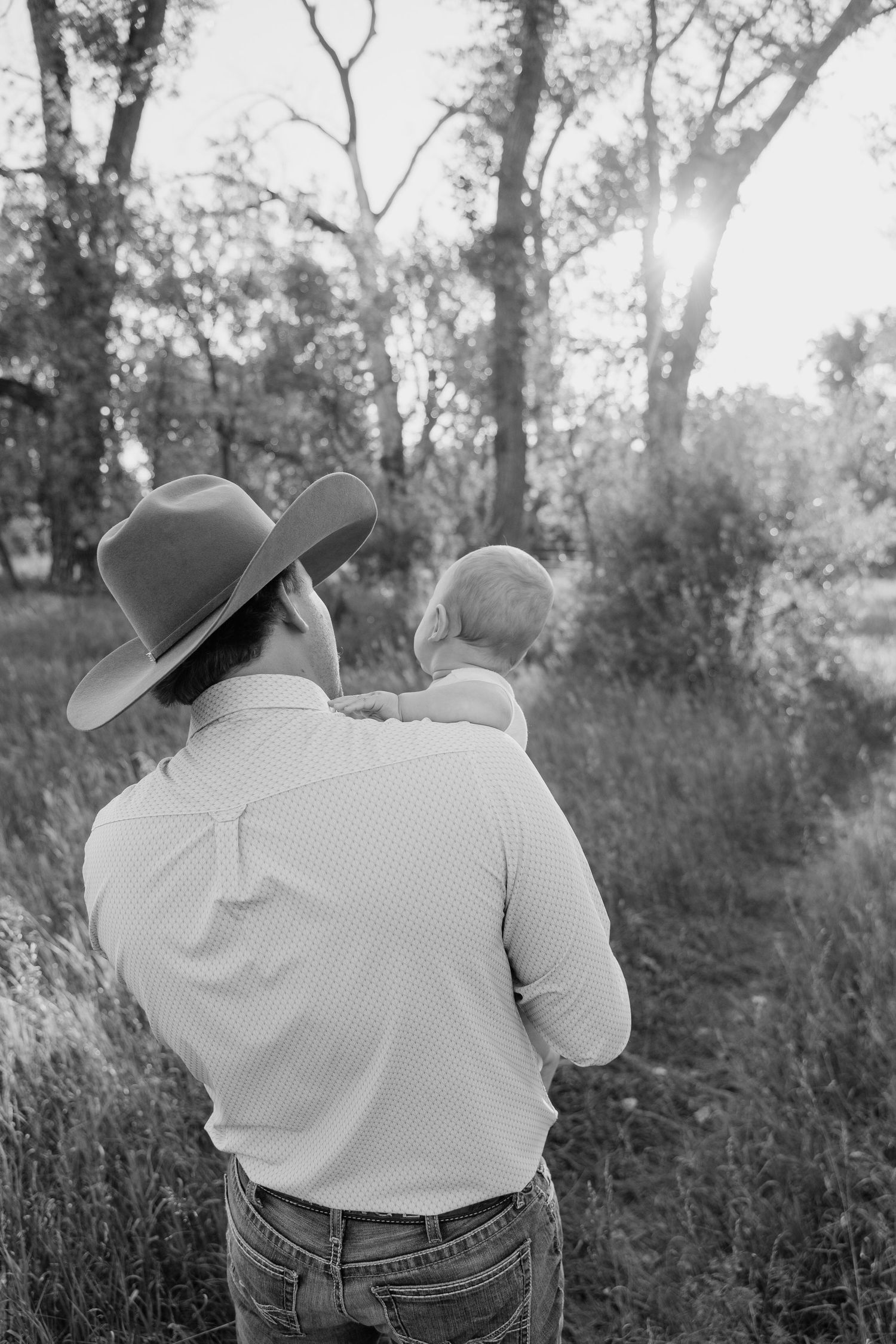 Black and white photo of a man in a cowboy hat holding a baby, standing in a field with trees and sunlight behind them.