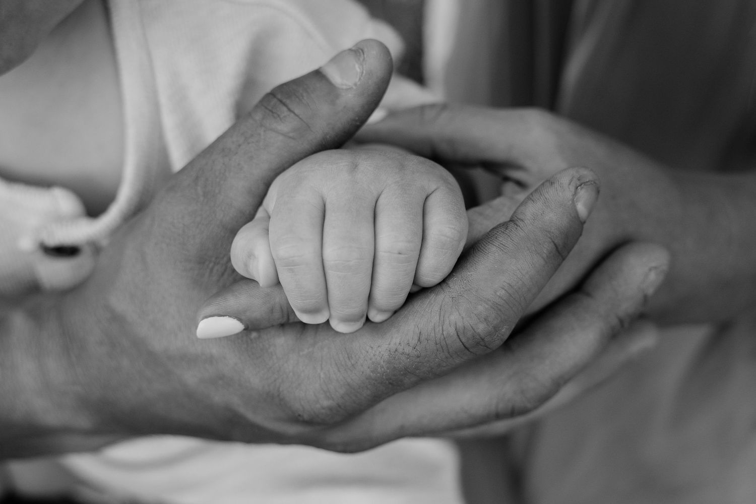 Black and white photo of a newborn baby's tiny hand cradled gently in two adult hands.