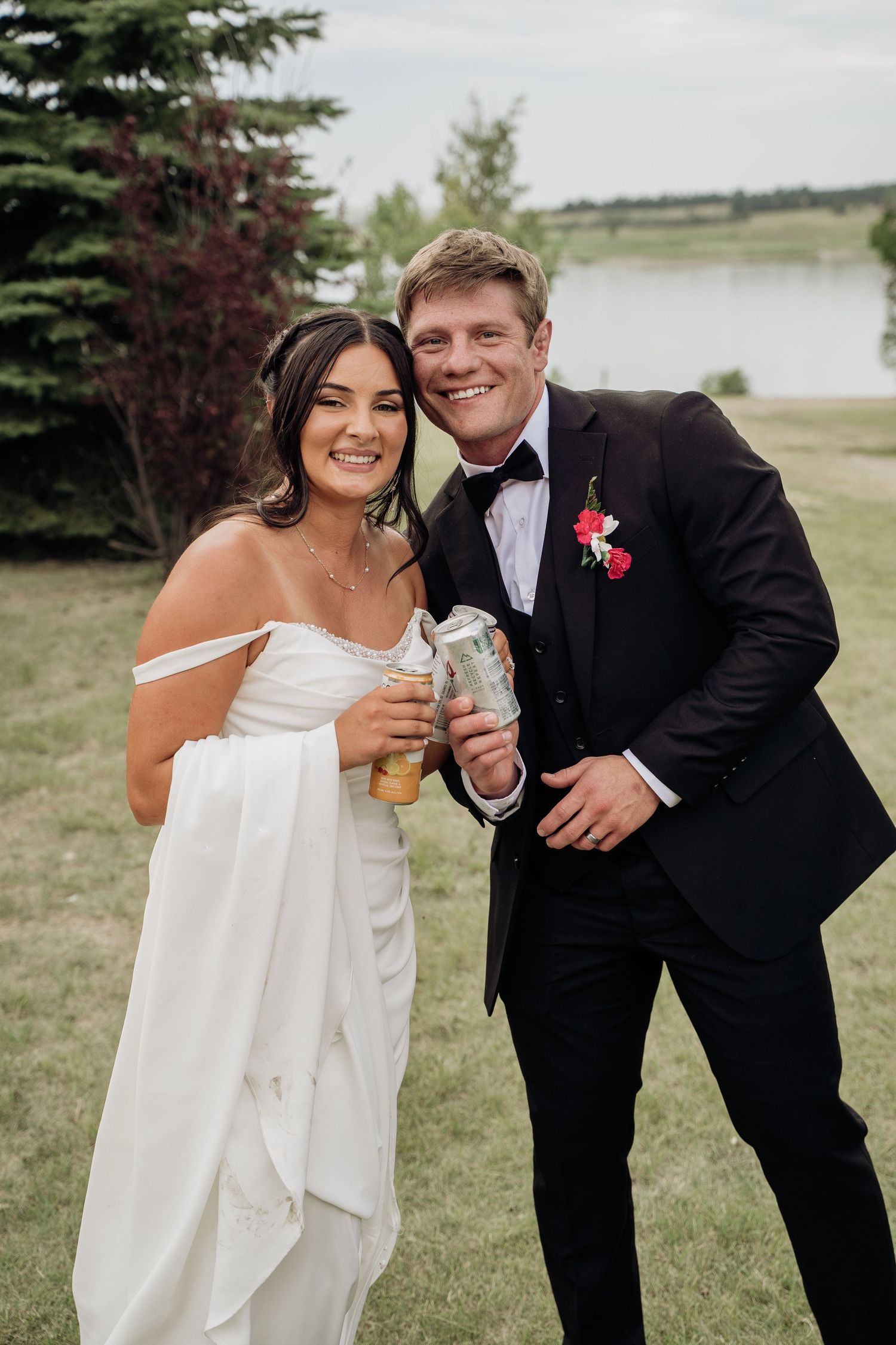 Smiling bride and groom holding drinks outdoors near lake after wedding ceremony.