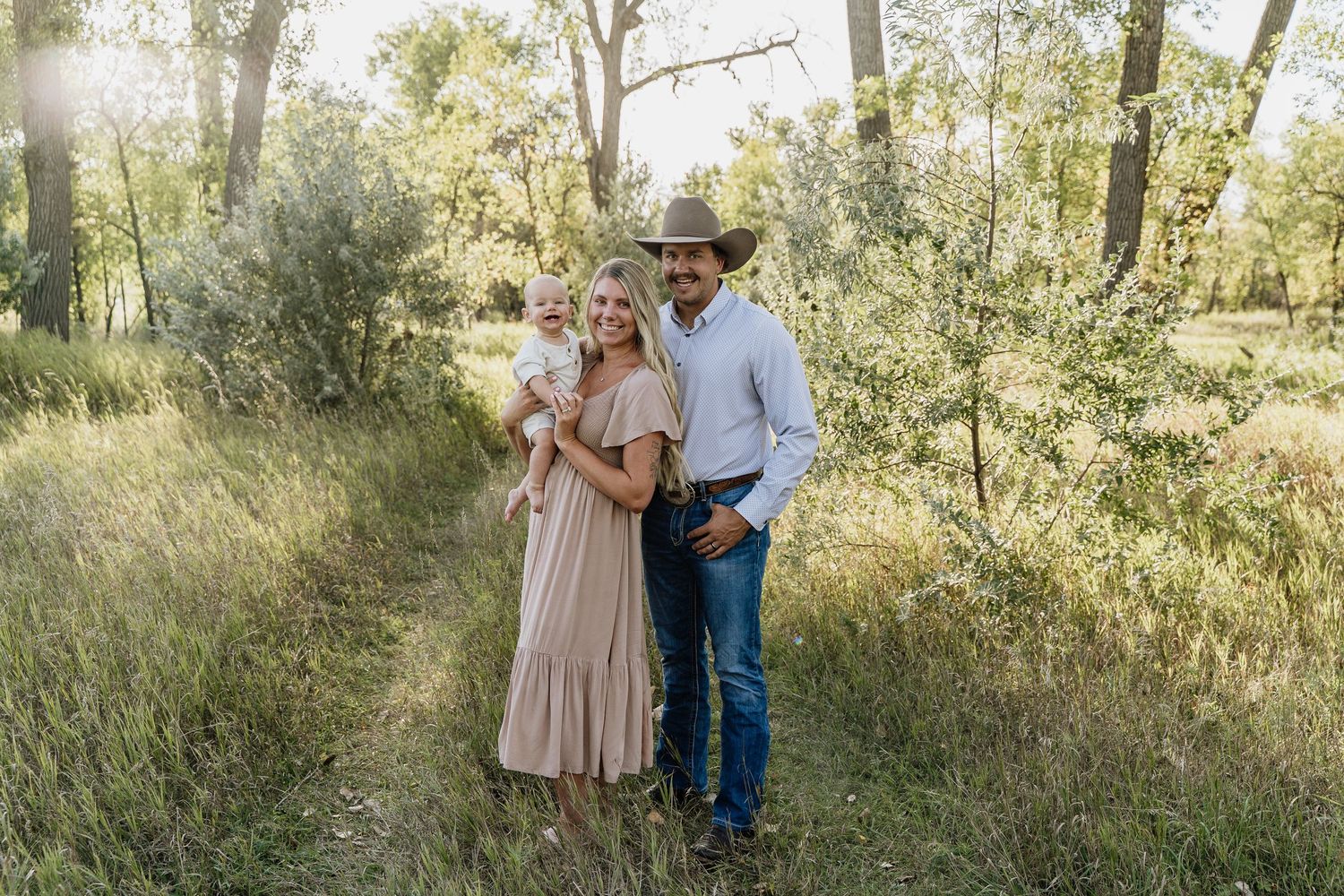 A young family poses outdoors in a lush green field, woman in beige dress holding baby, man in cowboy hat and jeans.