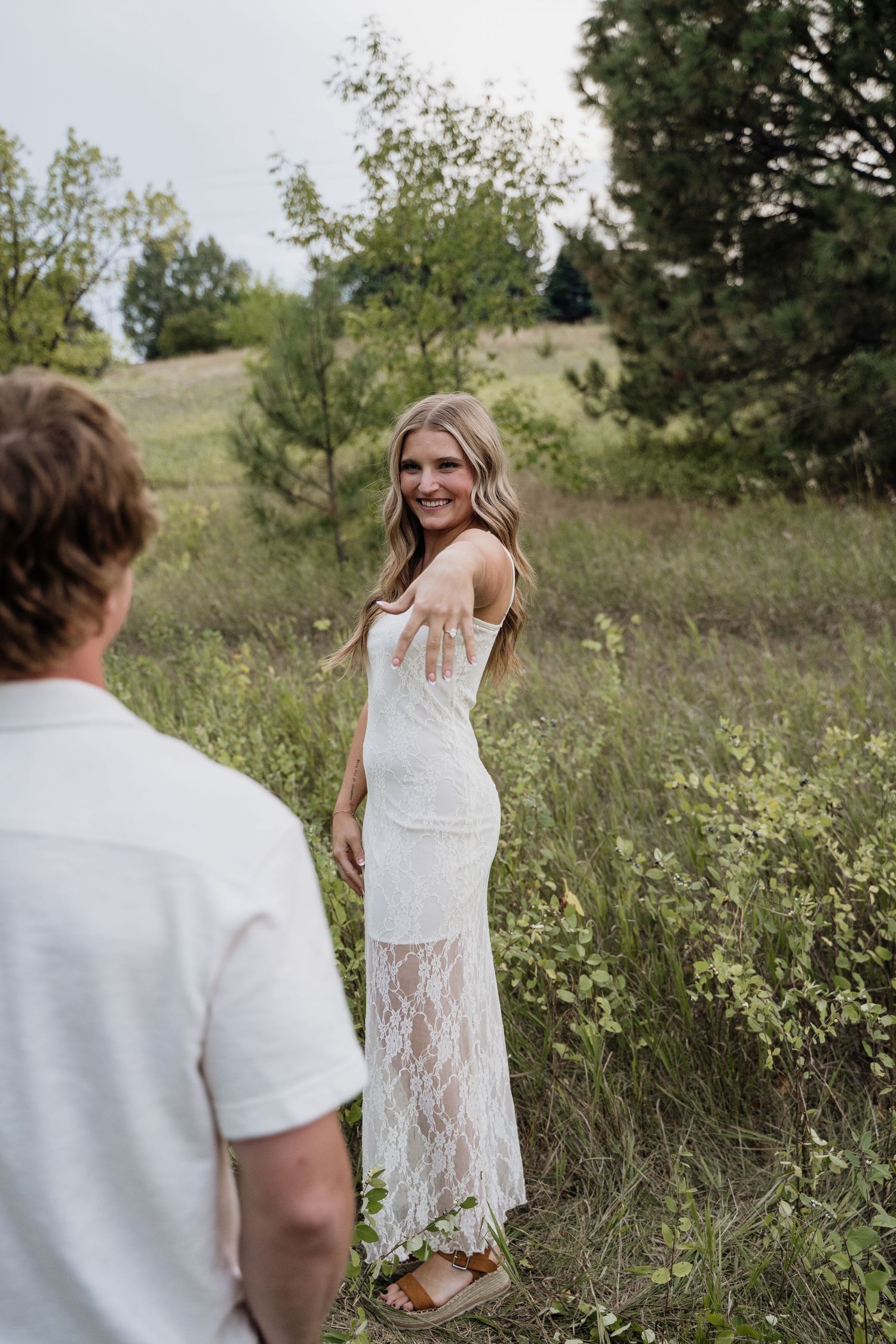 A smiling woman in a white lace dress reaches back toward a man in a grassy outdoor setting.