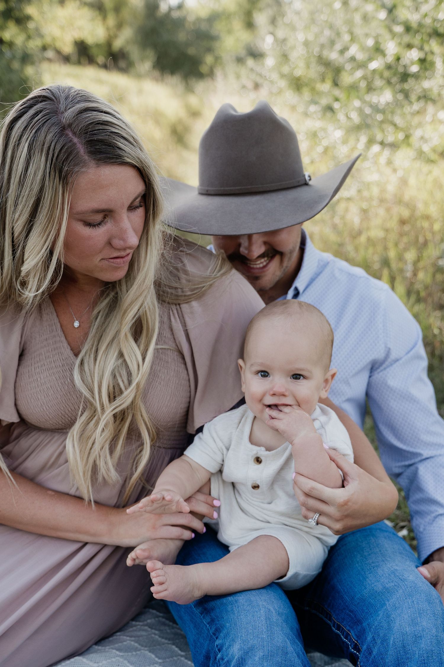 A smiling couple sits outdoors with their happy baby in white, mom in mauve dress, dad in cowboy hat and blue shirt.