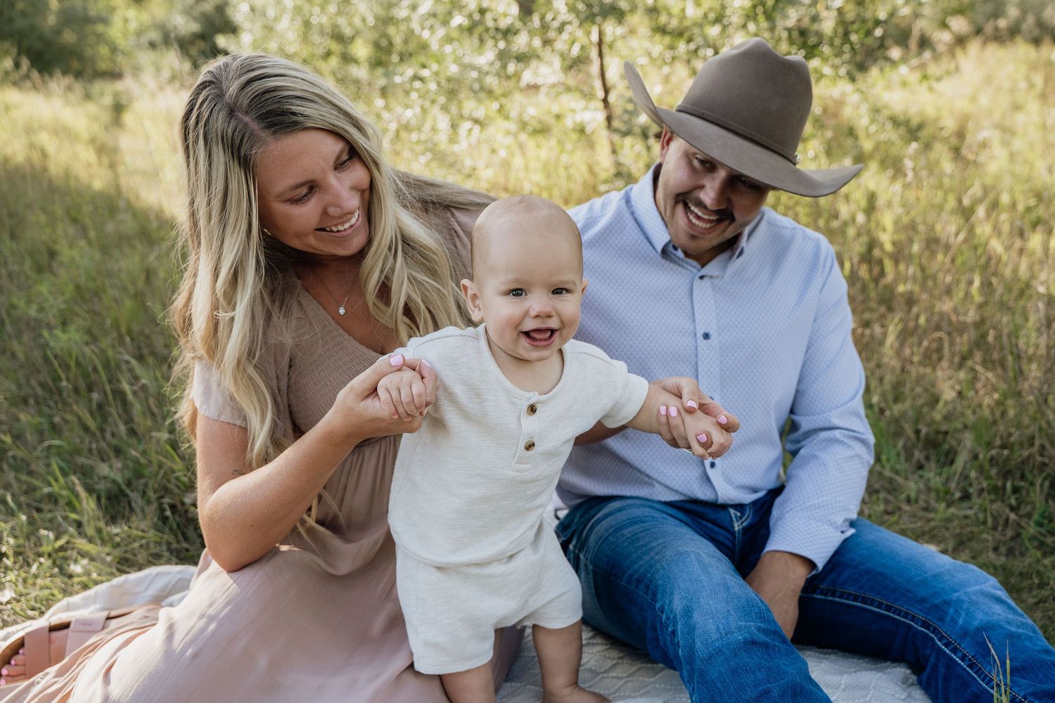 Happy family with smiling baby sitting outdoors in green field, parents holding toddler's hands in natural setting.