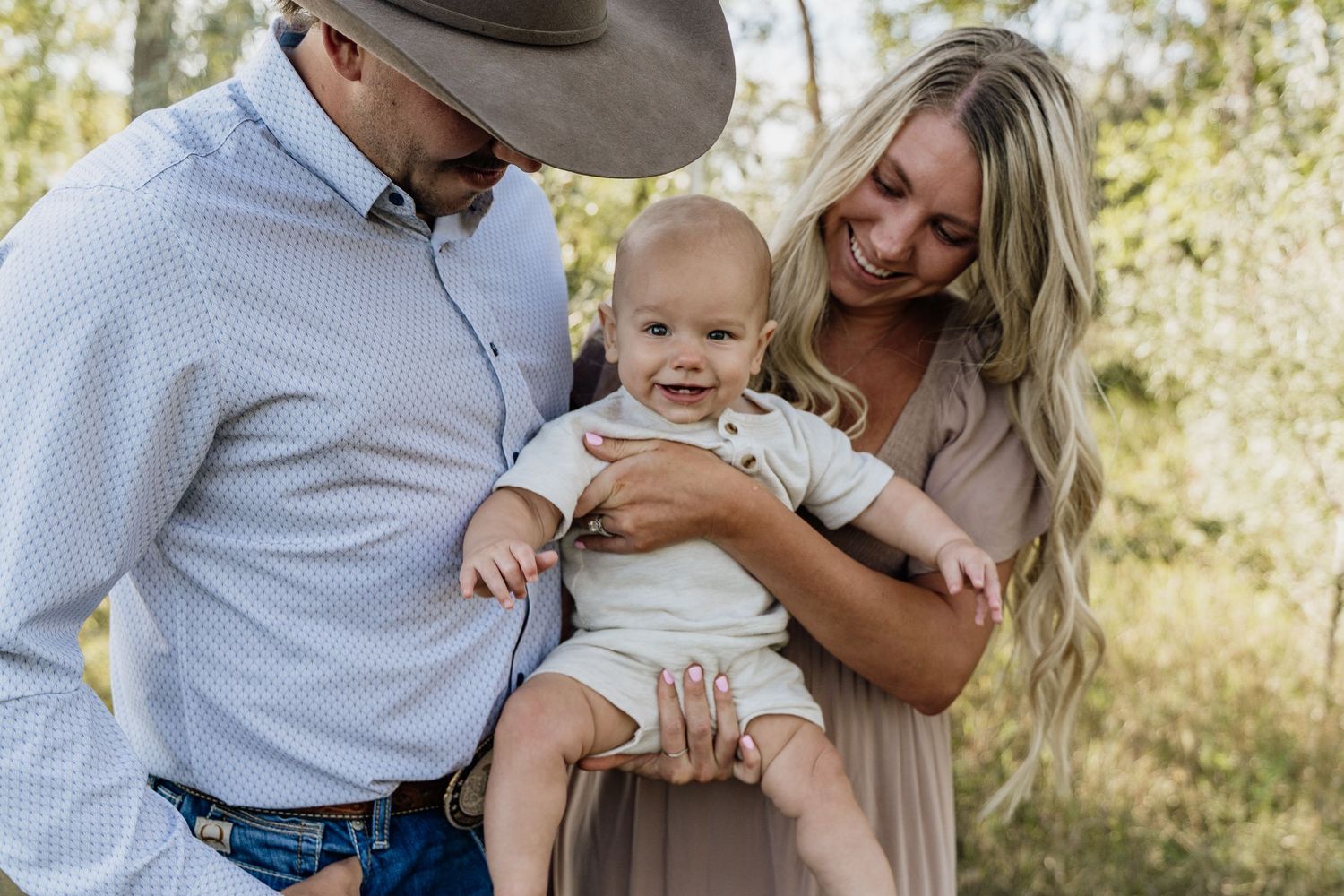 A smiling couple in a outdoor setting holds their happy baby, with the man wearing a cowboy hat and woman in a tan dress.