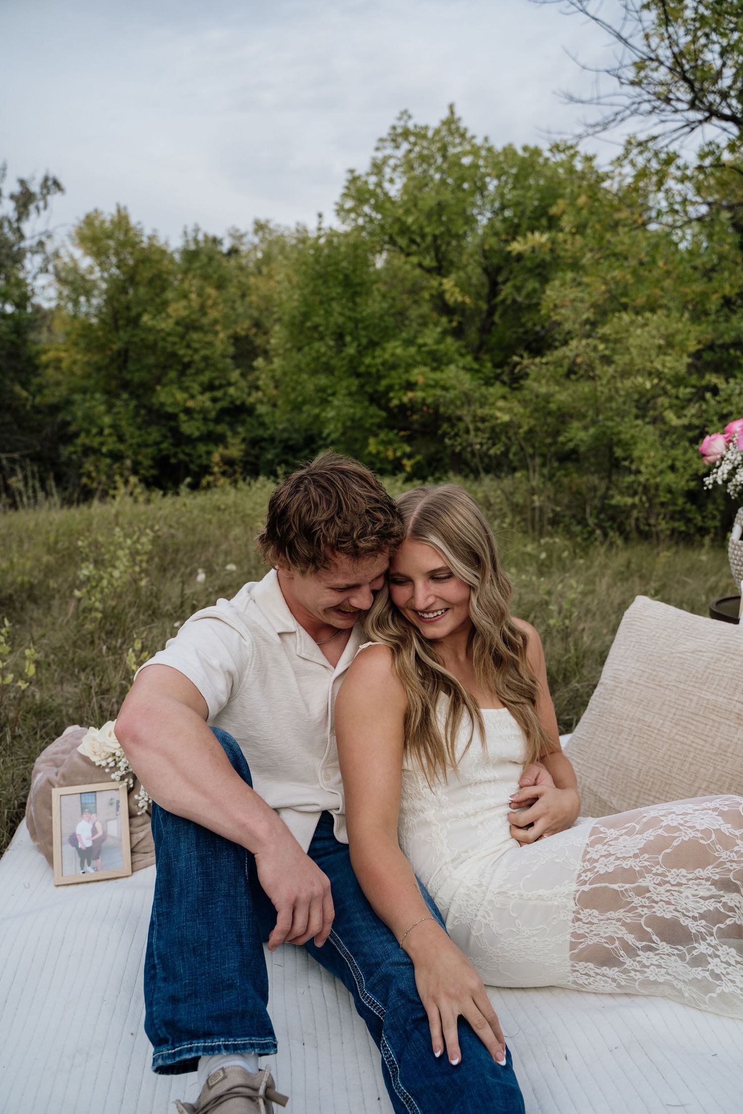 Couple sitting on white blanket in field, man embracing woman in lace dress, smiling together outdoors.