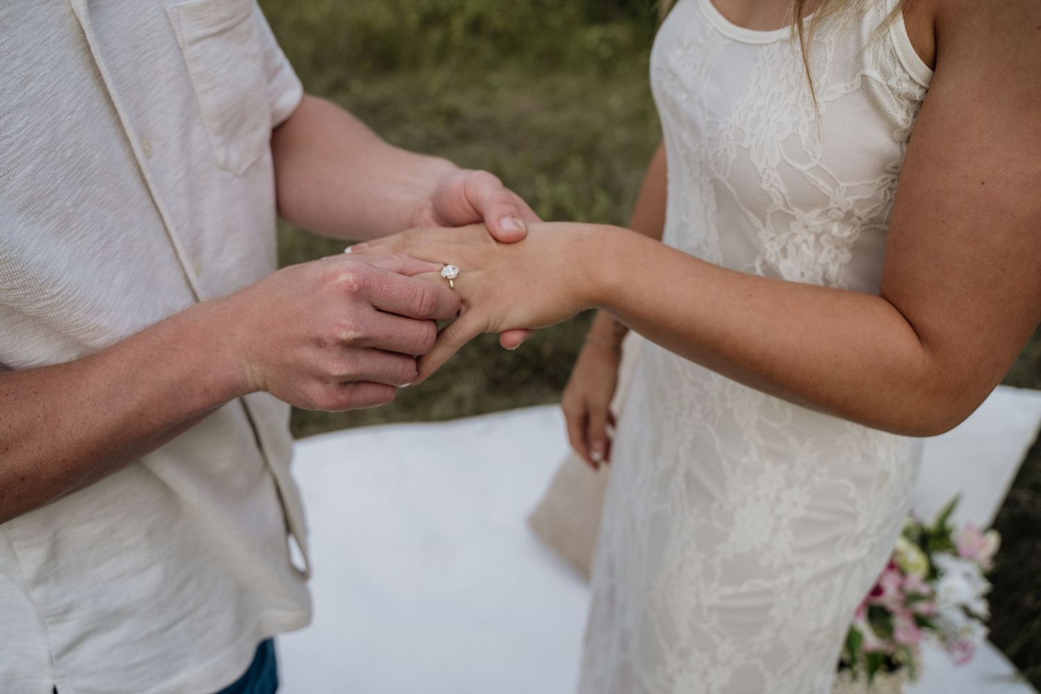 Groom placing a diamond engagement ring on bride's finger during outdoor wedding ceremony.