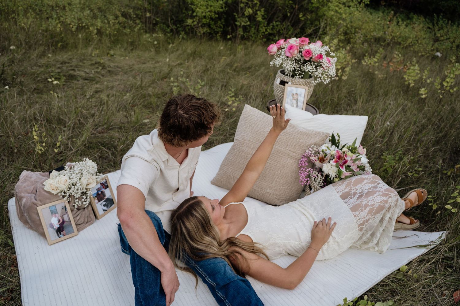 Couple relaxing on a white blanket outdoors with pink flowers and framed photos during a romantic picnic.