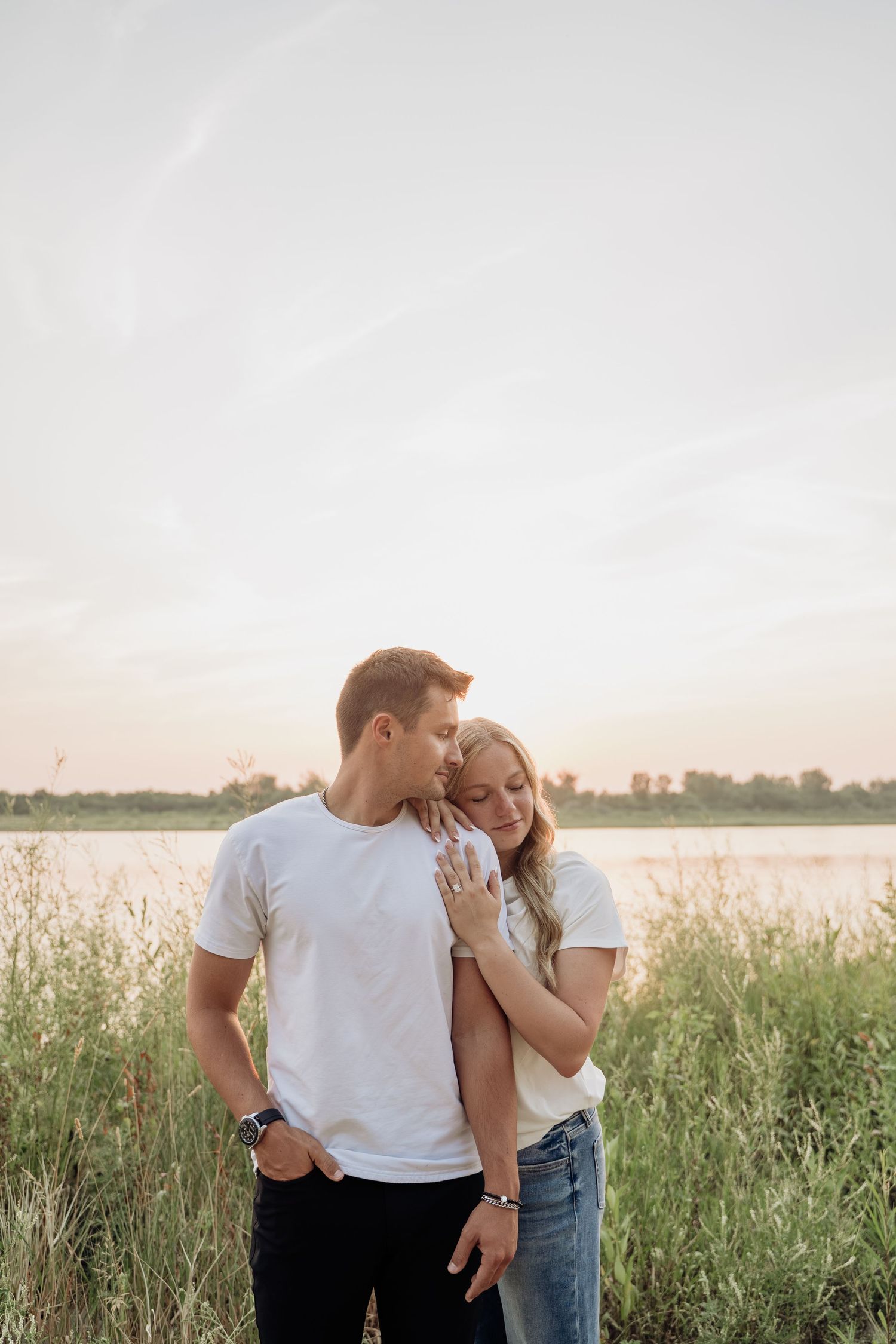 A couple shares a tender moment near a lake at dusk, woman resting on mans chest.