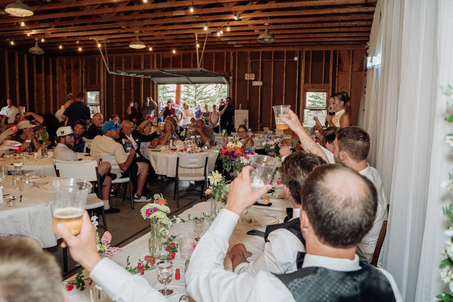 Wedding guests raise glasses in a toast inside a rustic barn decorated with colorful floral centerpieces.