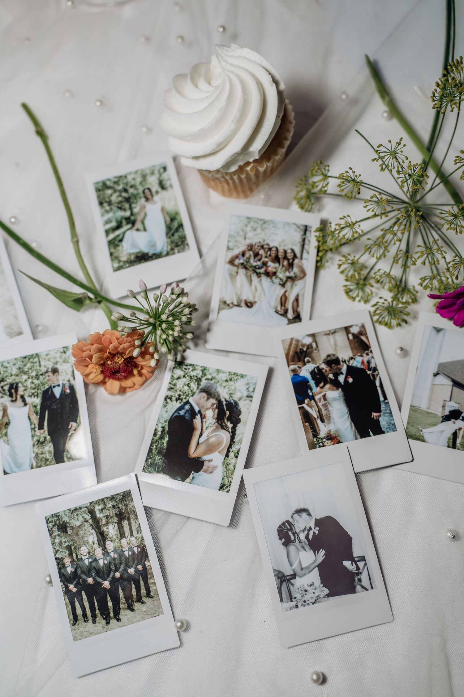 Polaroid wedding photos scattered on white fabric with a cupcake, flowers, and pearl decorations.