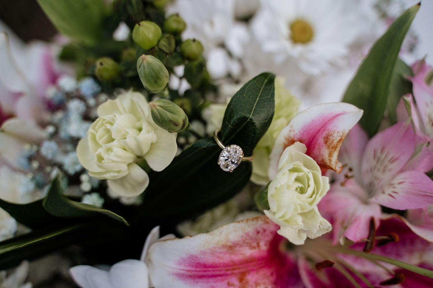 Diamond engagement ring resting on a bridal bouquet of pink lilies, white roses, and green foliage.