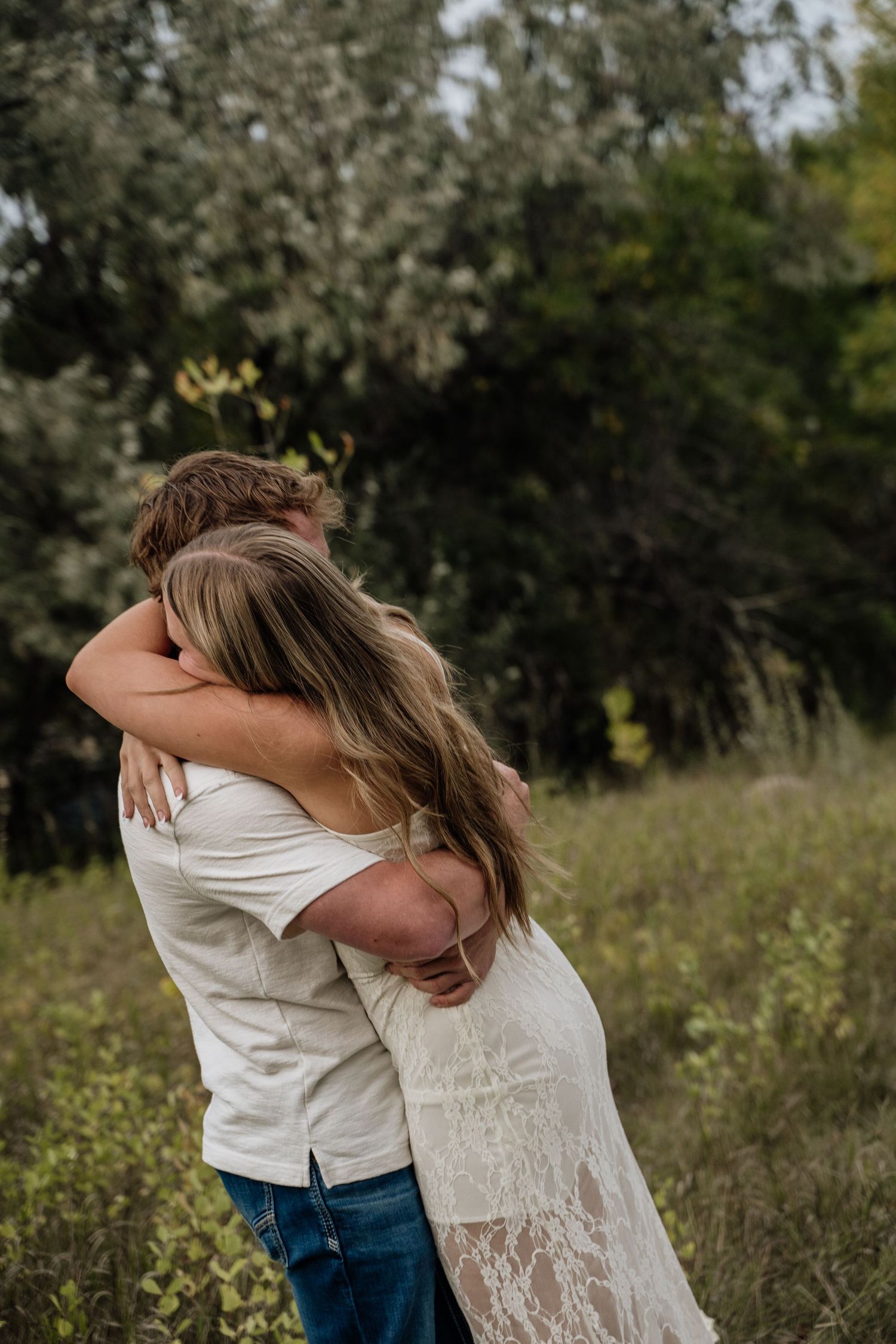 Couple embracing in a lush green field, woman in lace dress and man in white shirt and jeans, romantic outdoor portrait.