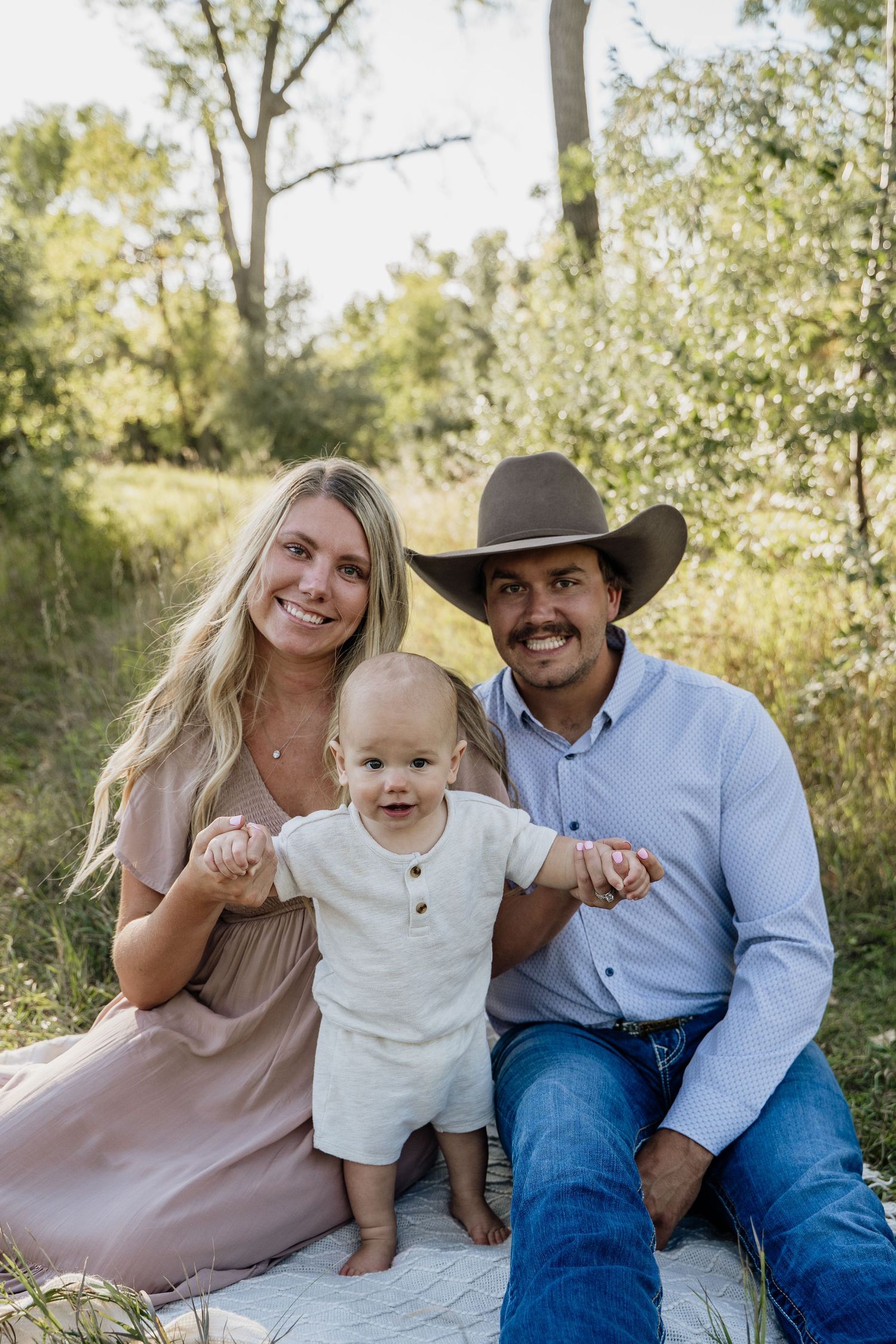 A smiling family of three poses outdoors, with a blonde woman, a man in a cowboy hat, and a baby standing between them.