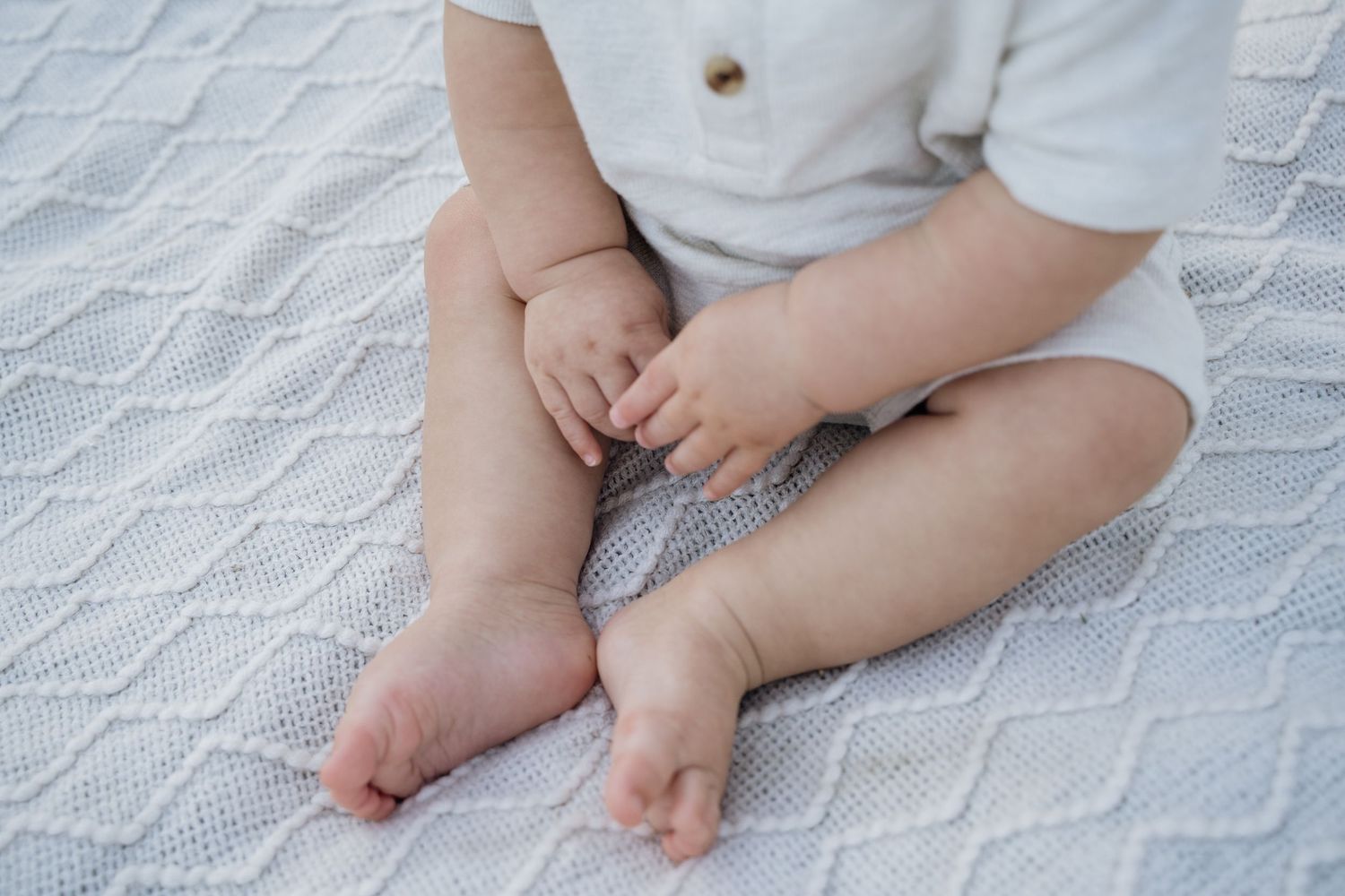 Close-up of a baby sitting cross-legged on a white knit blanket, wearing a light linen outfit.