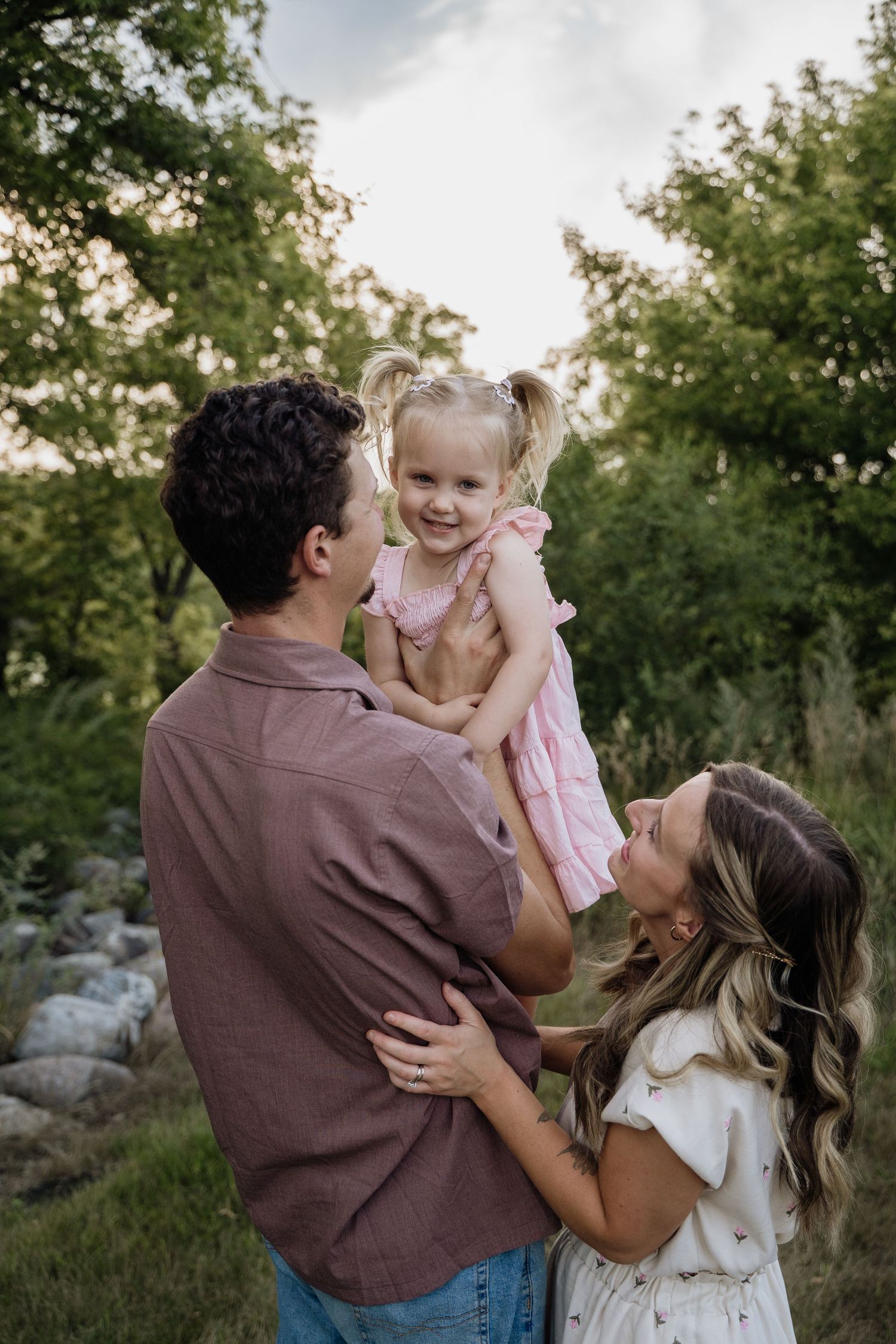 Young family with toddler girl in pink dress being lifted up outdoors among green trees.
