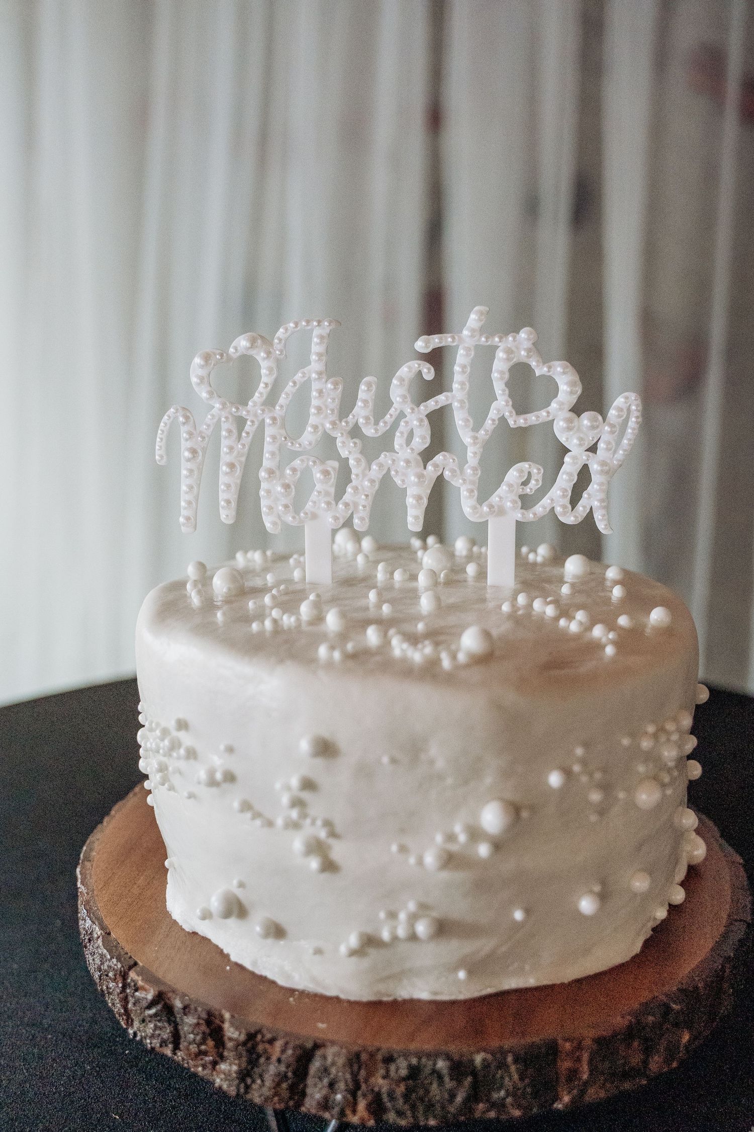 Elegant single-tier white wedding cake with pearl decorations and a glittery Just Married topper on a wood slice.