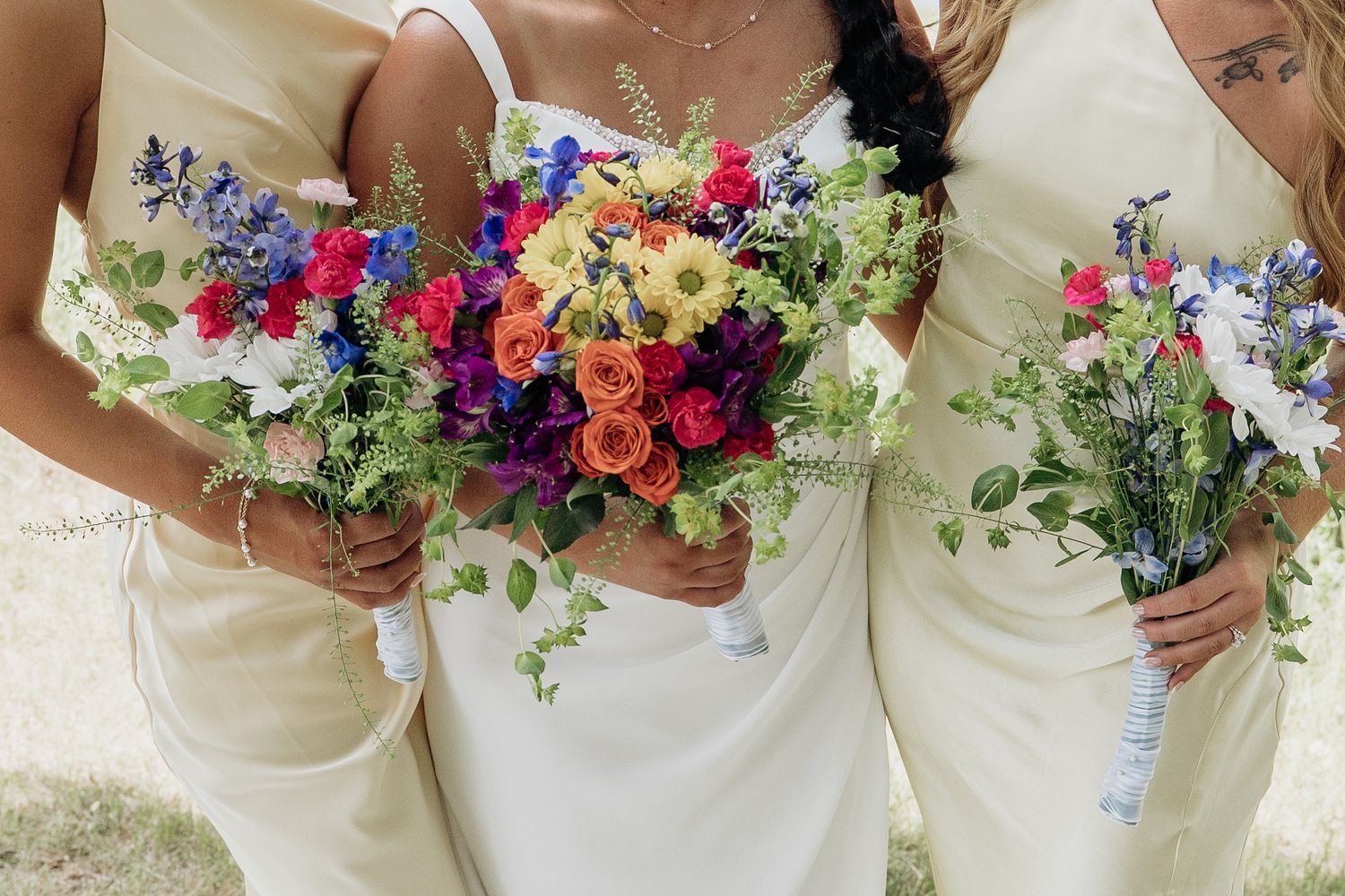 Bride in white gown and bridesmaids in cream dresses holding colorful wildflower bouquets.